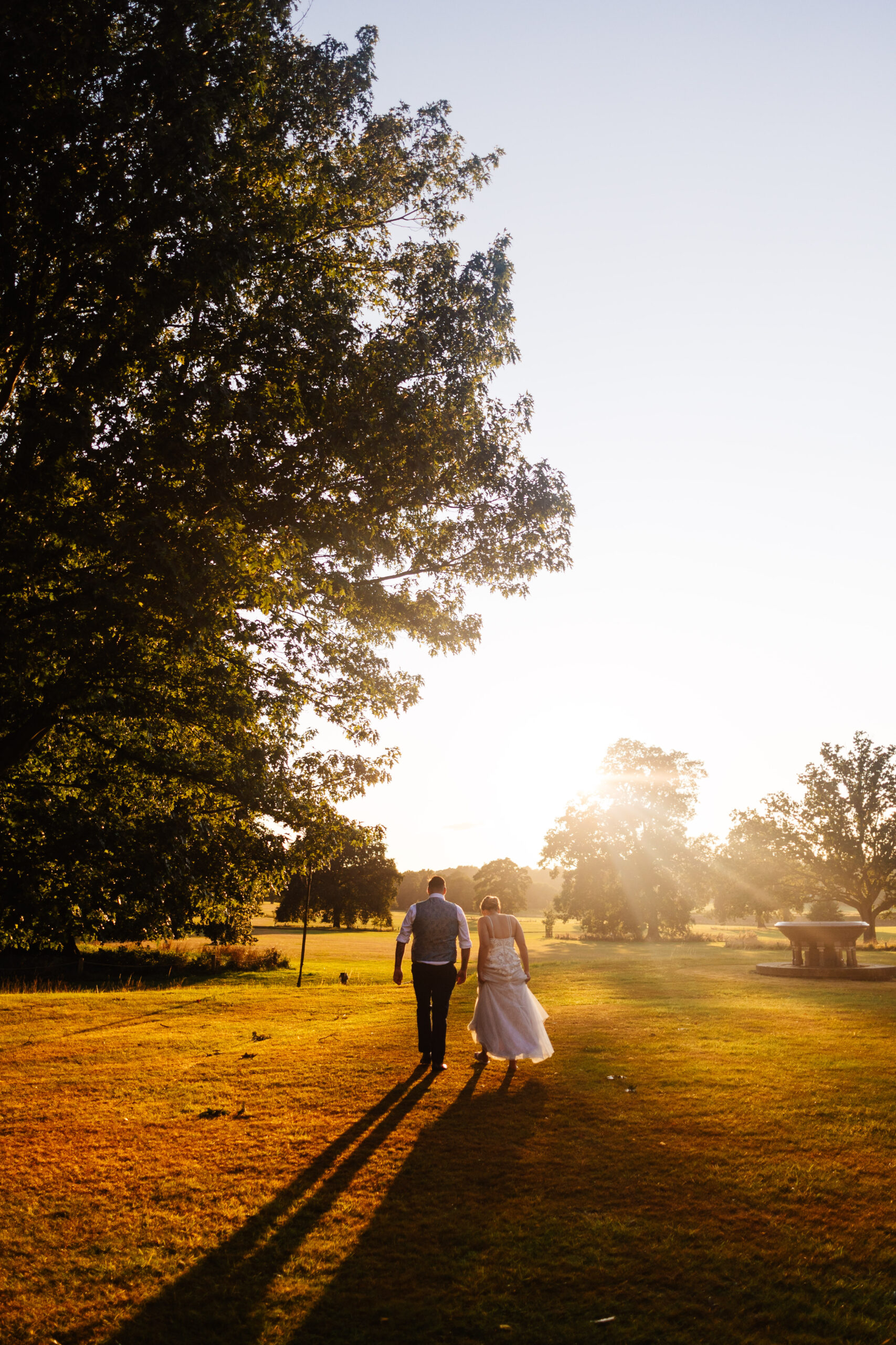 A photo of the bride and groom outside. The sun is pouring through the trees. They have their backs to the camera and are walking in to the distance.