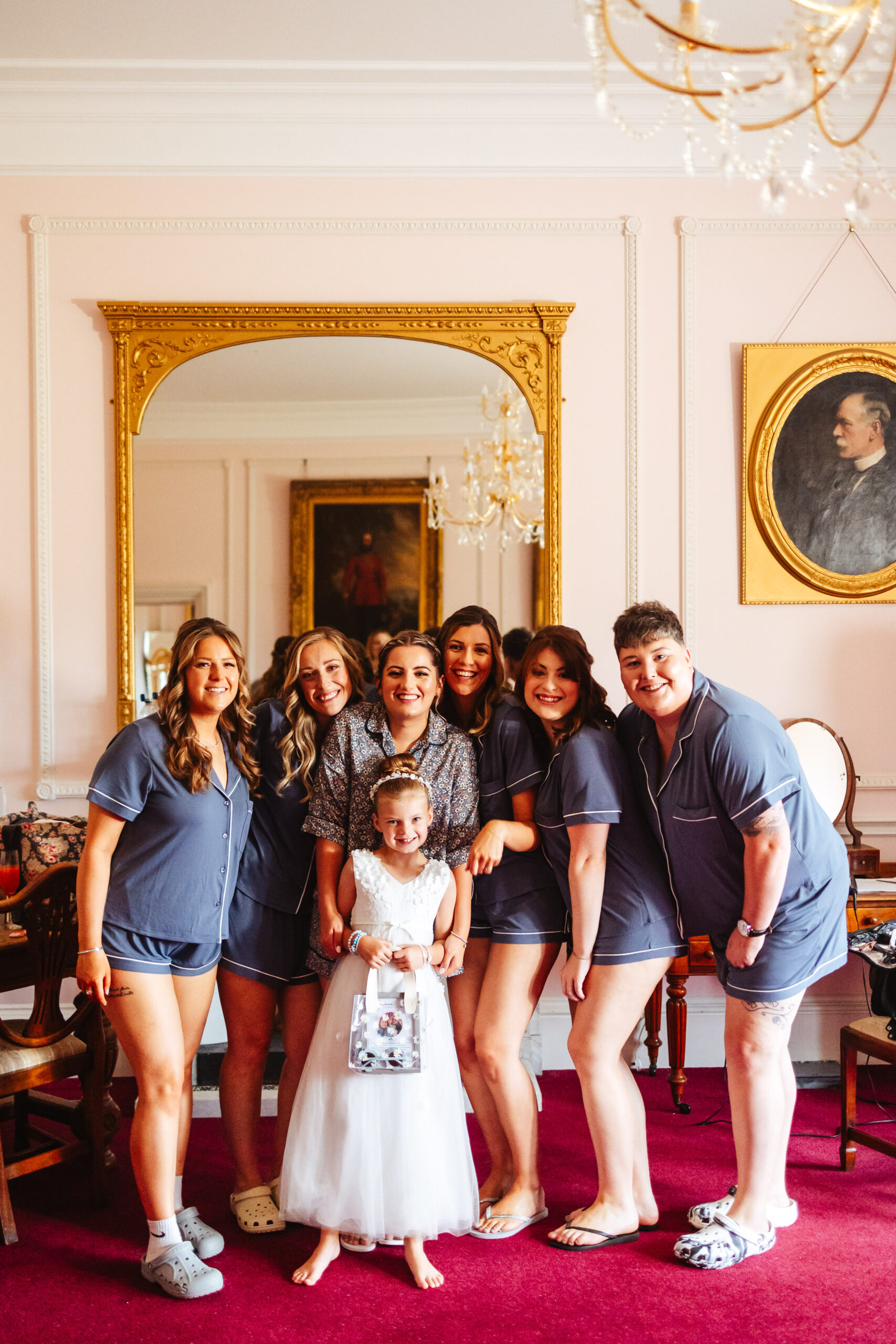 The bride and her bridesmaids and flower girl. They are all huddled around and are smiling at the camera. The bridesmaids are in their pyjamas and the flower girl is in her white dress but has no shoes on.