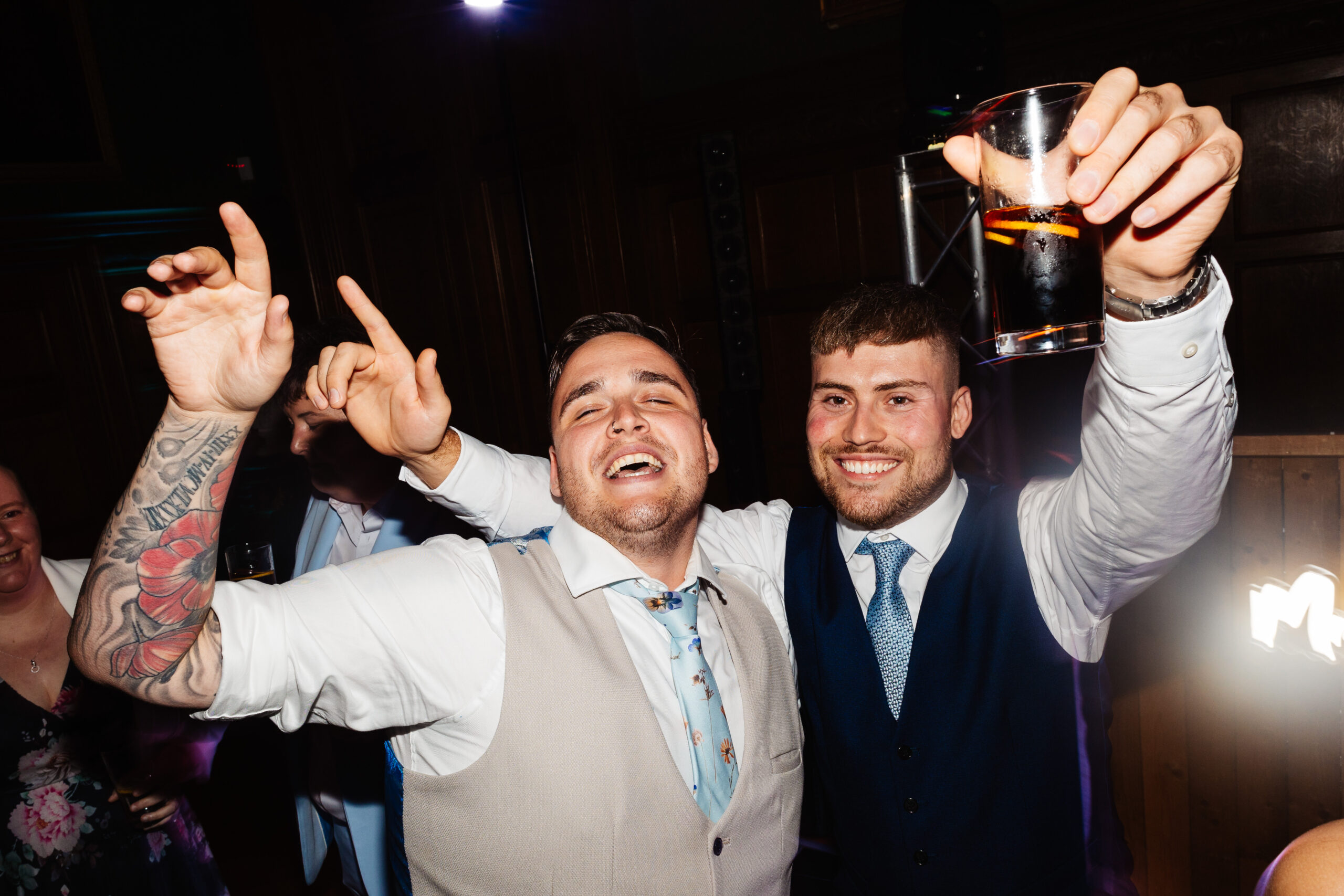 The groom and a guest. They both have one hand in the air, dancing and the guest has a drink in the other hand.