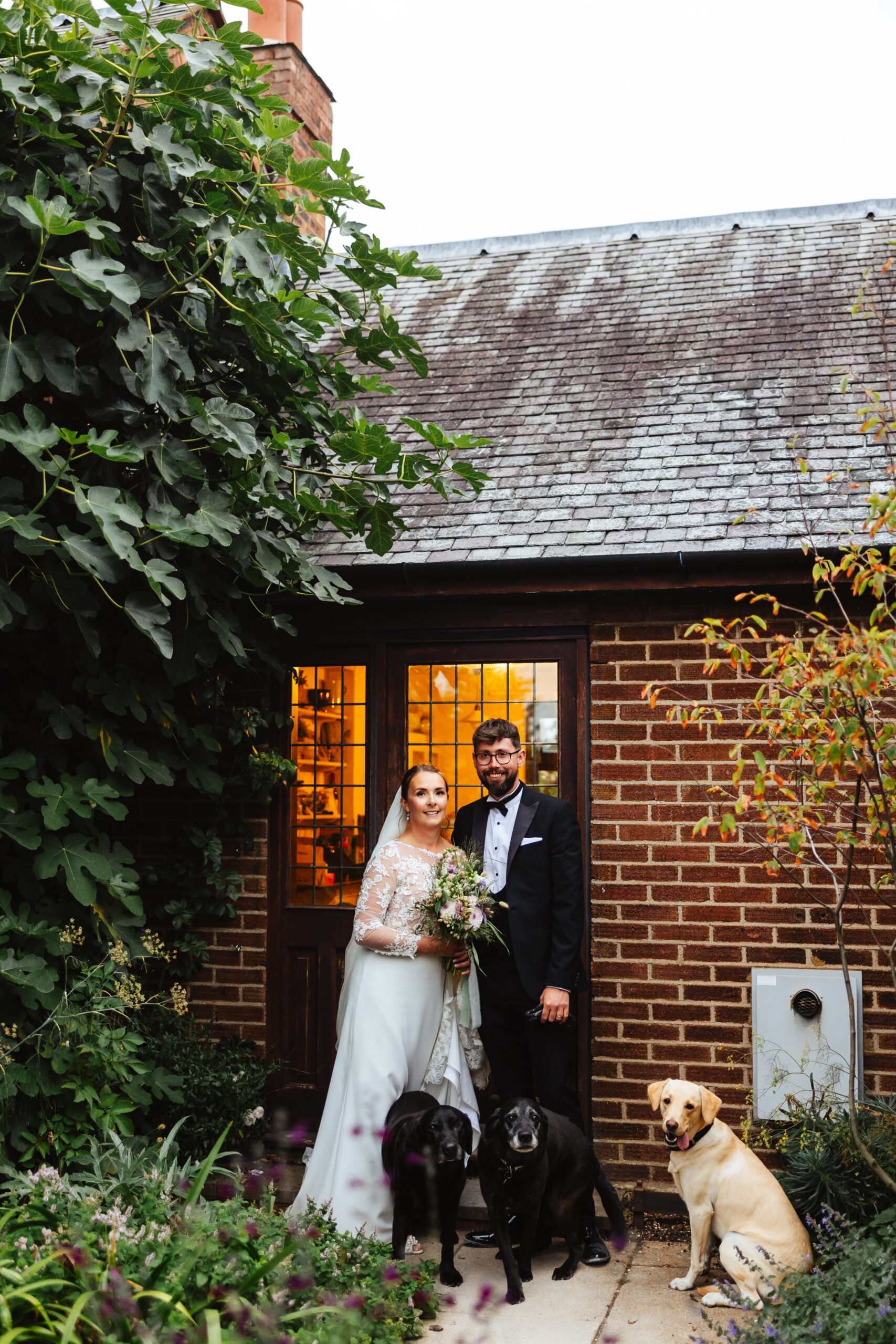 The bride and groom stood in front of their house. There are three dogs by their feet - two black labradors and a golden one. The golden labrador has its tongue out.