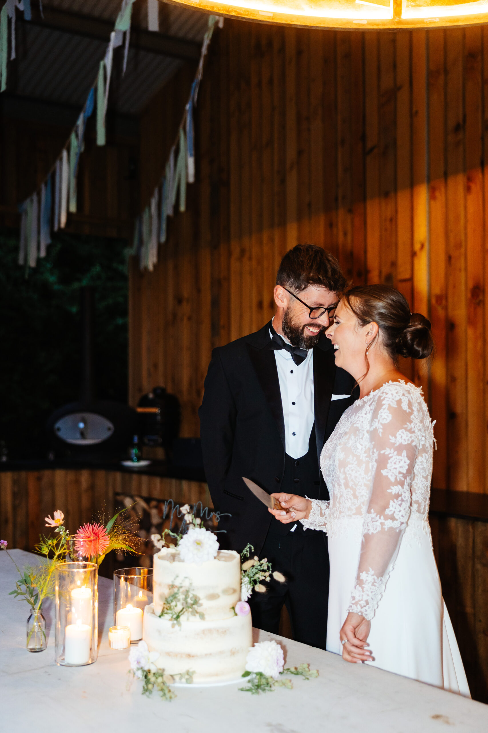 A photo of the bride and groom cutting their cake. The cake is a two tiered, naked cake with soft beieg icing on it and florals scattered on it. There is a Mr & Mrs sign on top. The couple are looking at each other and smiling and the bride has a knife in her left hand ready to cute the cake.