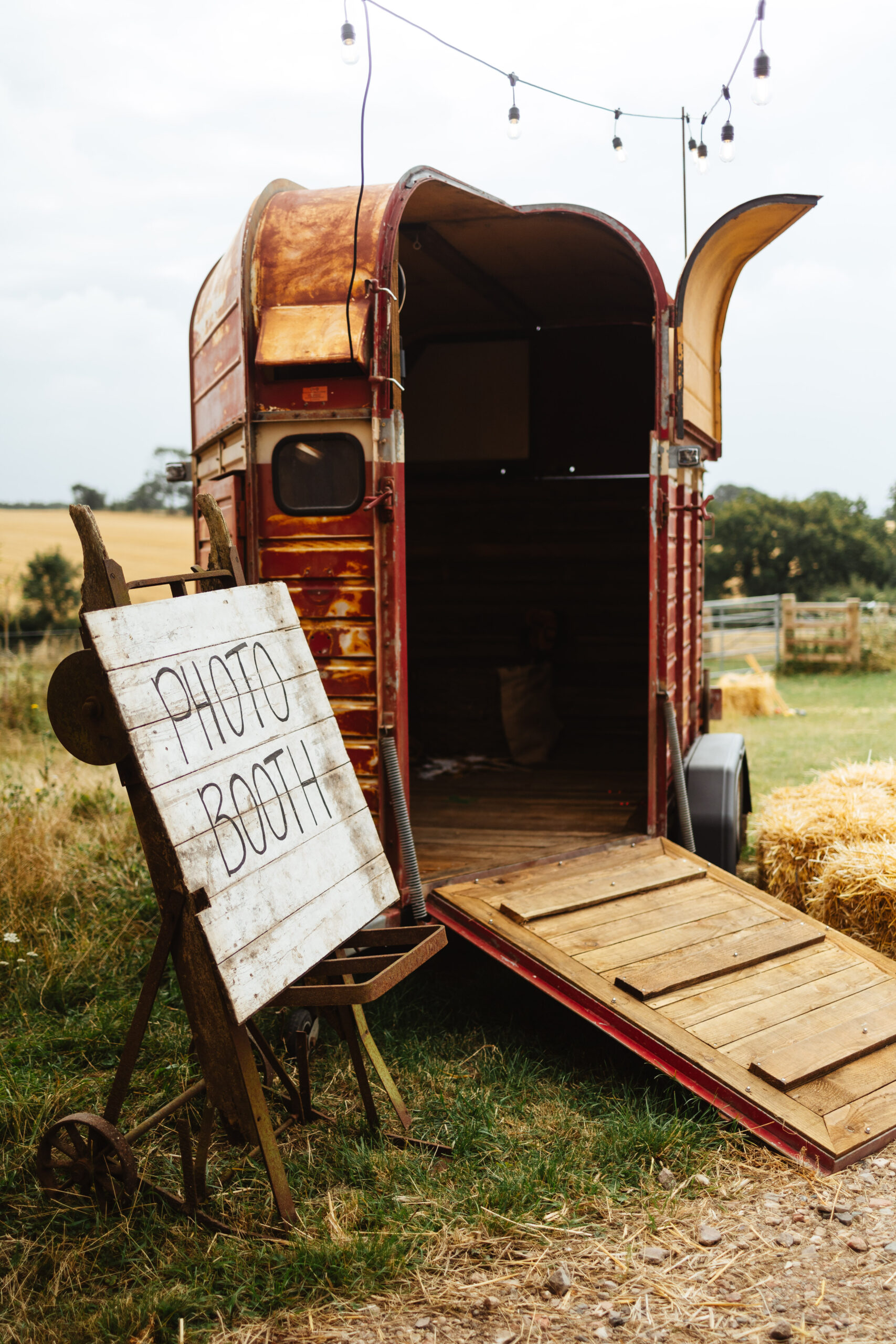 A horse box that has been converted in to a photo booth. There is a wooden sign that says Photo Booth on it. This is in a field.