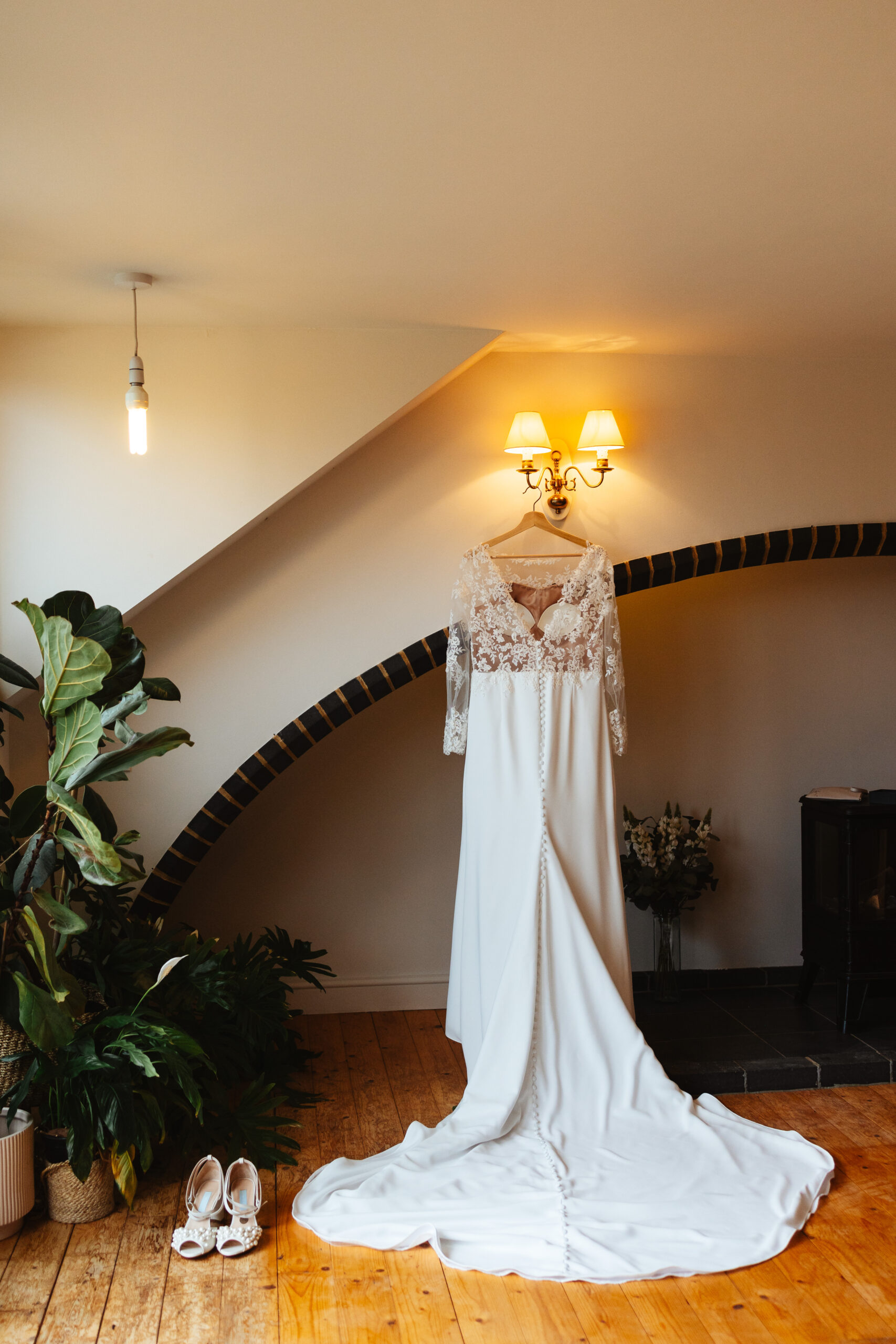 The bride's dress hanging up inside. It is a white dress with a lace top and sleeves. There are small buttons down the front. It is a long dress with a long train.