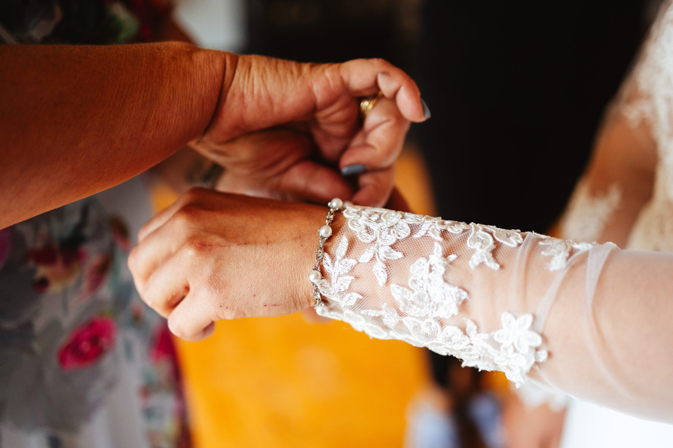 The bride's arm. She is wearing her wedding dress so you can see her sleeve detail. Her mum is helping to put on her bracelet.