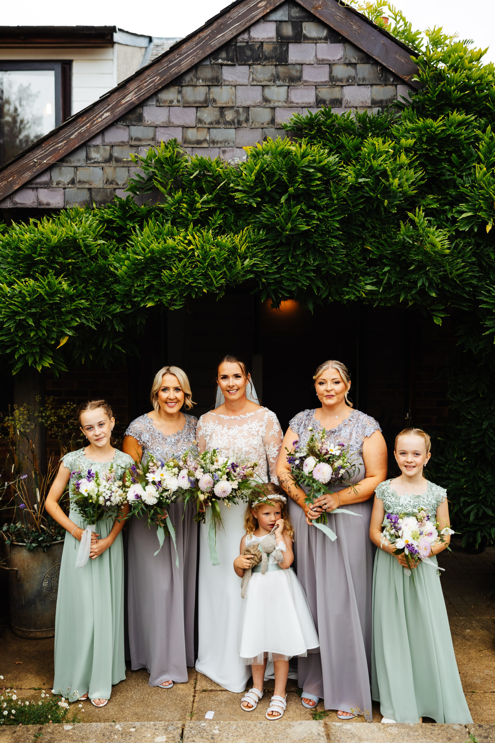 The bride, bridesmaids and flower girls standing outside. The bridesmaids are in a dull lilac colour dress and the flower girls are in a turquoise dress. They are all long and flowing. The little flower girl is in white and is holding her little rabbit.