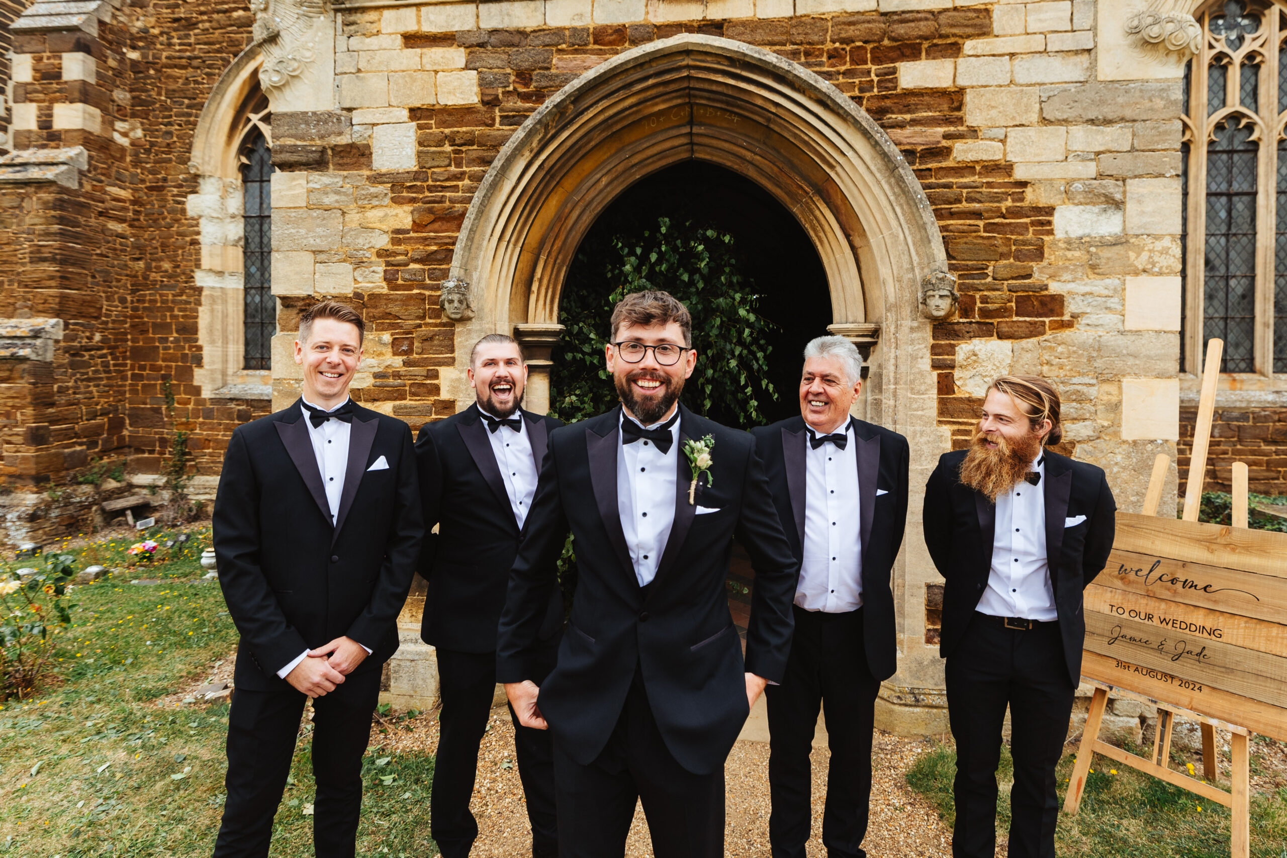 The groom and his groomsmen in their tux outside the church. They are looking at the groom and at the camera and are all smiling.