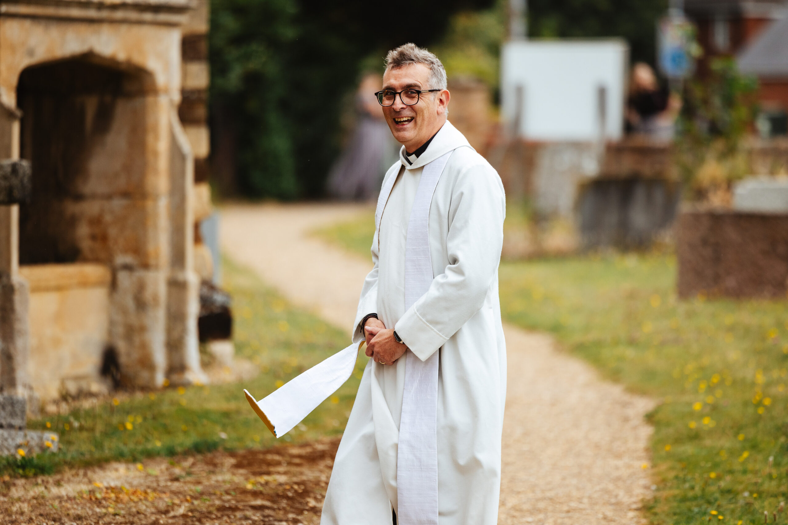 The priest outside the church. He is wearing white and is looking at the camera and smiling.