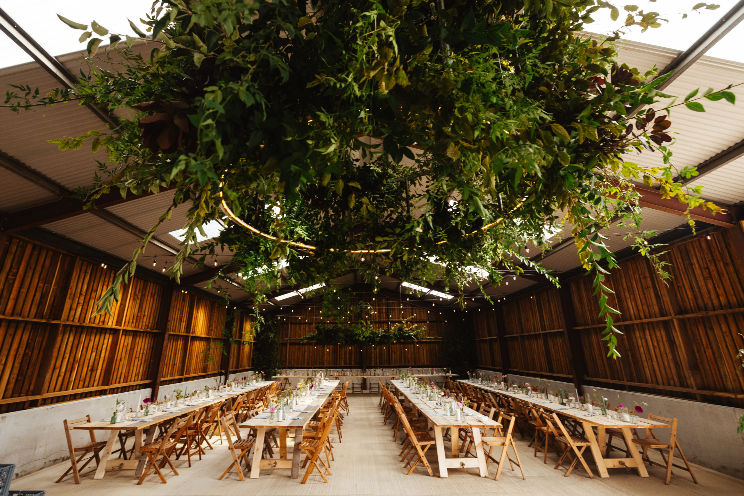 A large barn with long tables running in the middle of it. There is a large circle of greenery hanging above the table.