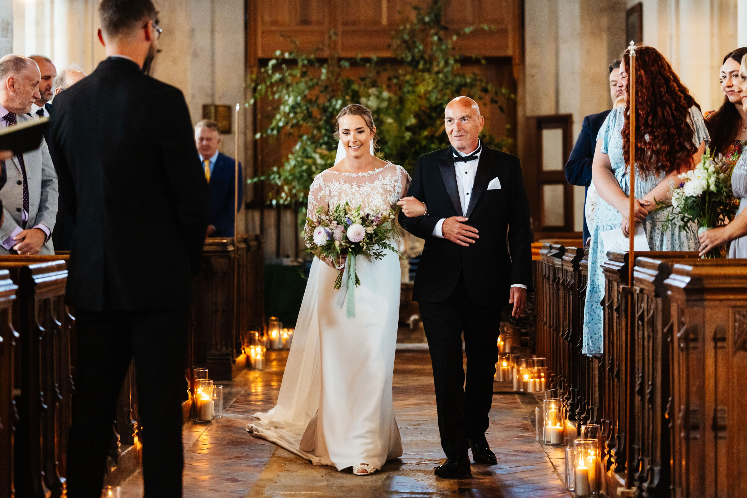 The bride and her father walking down the aisle. They are in a traditional church and her guests are in the pews. They are all looking at the bride and smiling. Her groom is at the front of the aisle and is looking at her smiling.