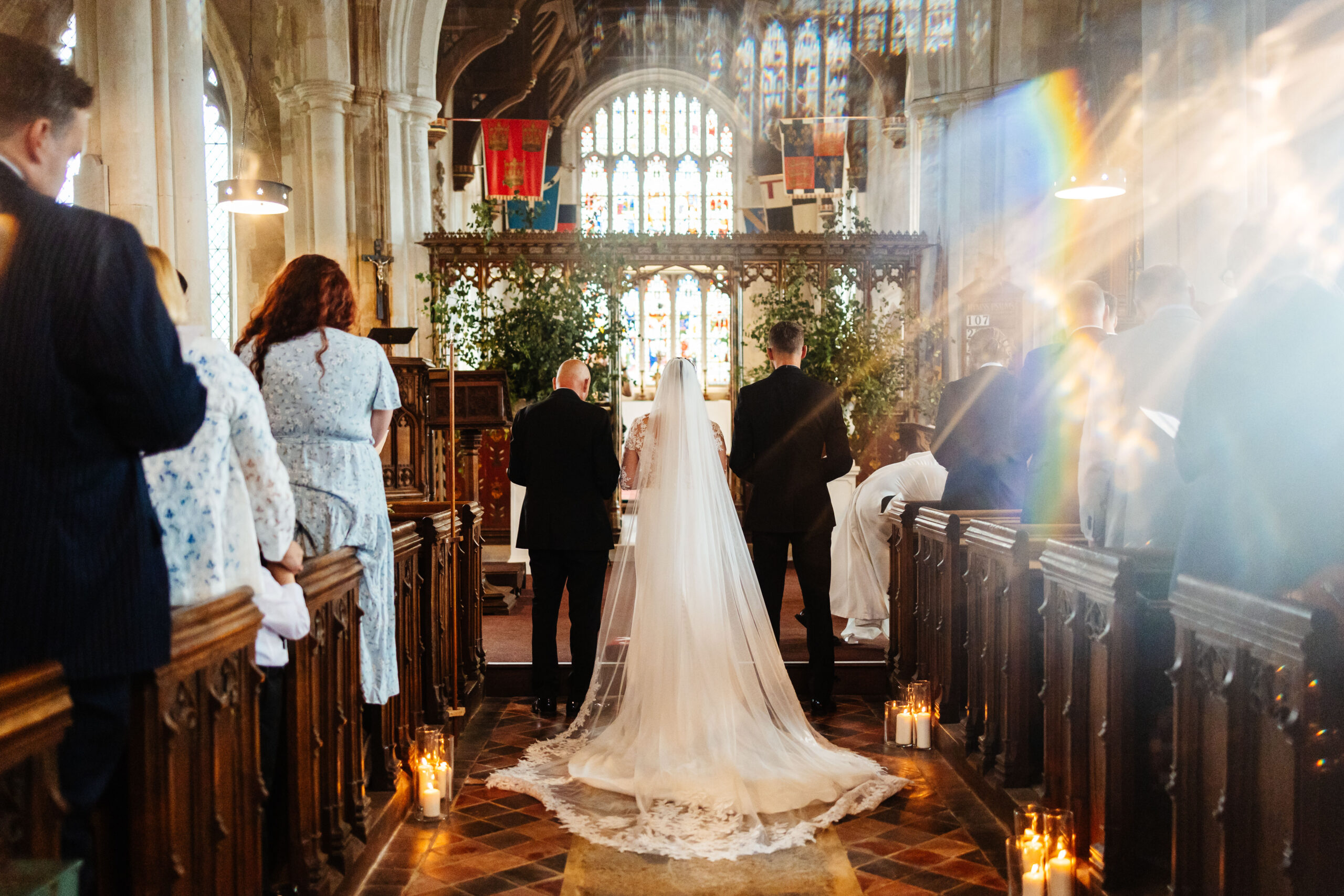 A stunning photo of the back of the bride and groom. The sun is shining through the stain glass window and it is creating a rainbow effect through the glass. You can see the bride's train of the dress and veil on the floor.