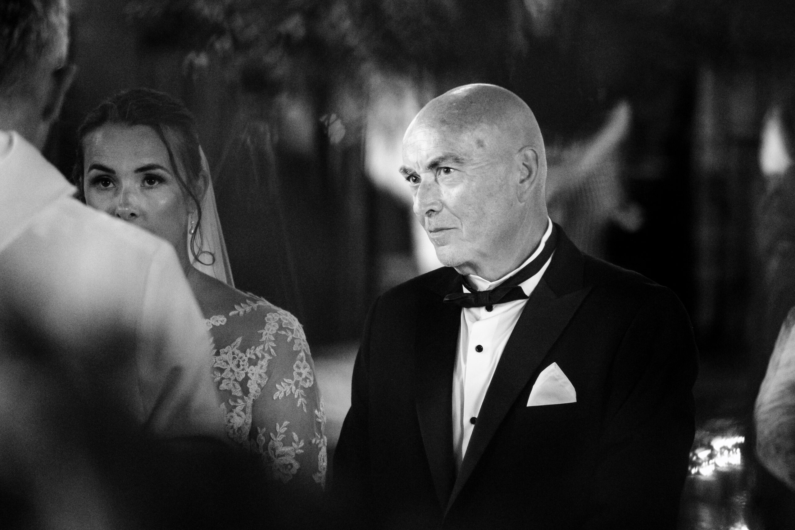 A black and white image of the father of the bride. He is looking at the priest and you can see his daughter (the bride) also in shot.