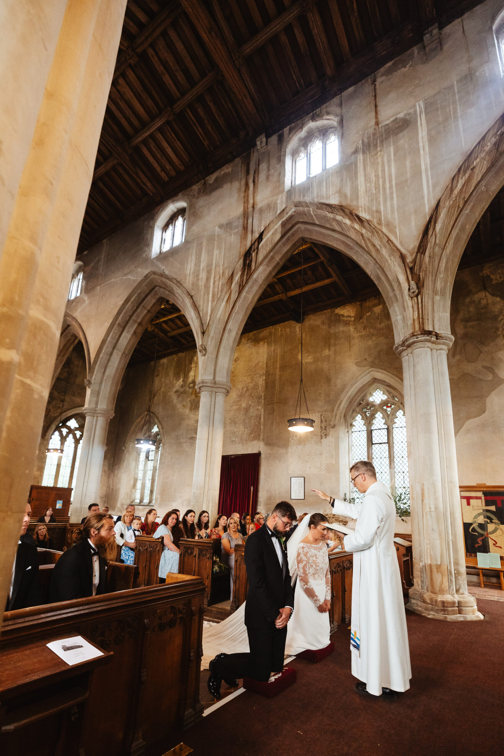 The bride and groom are on their knees. The priest is blessing them, holding a book in one hand and his hand over the couple in the other.