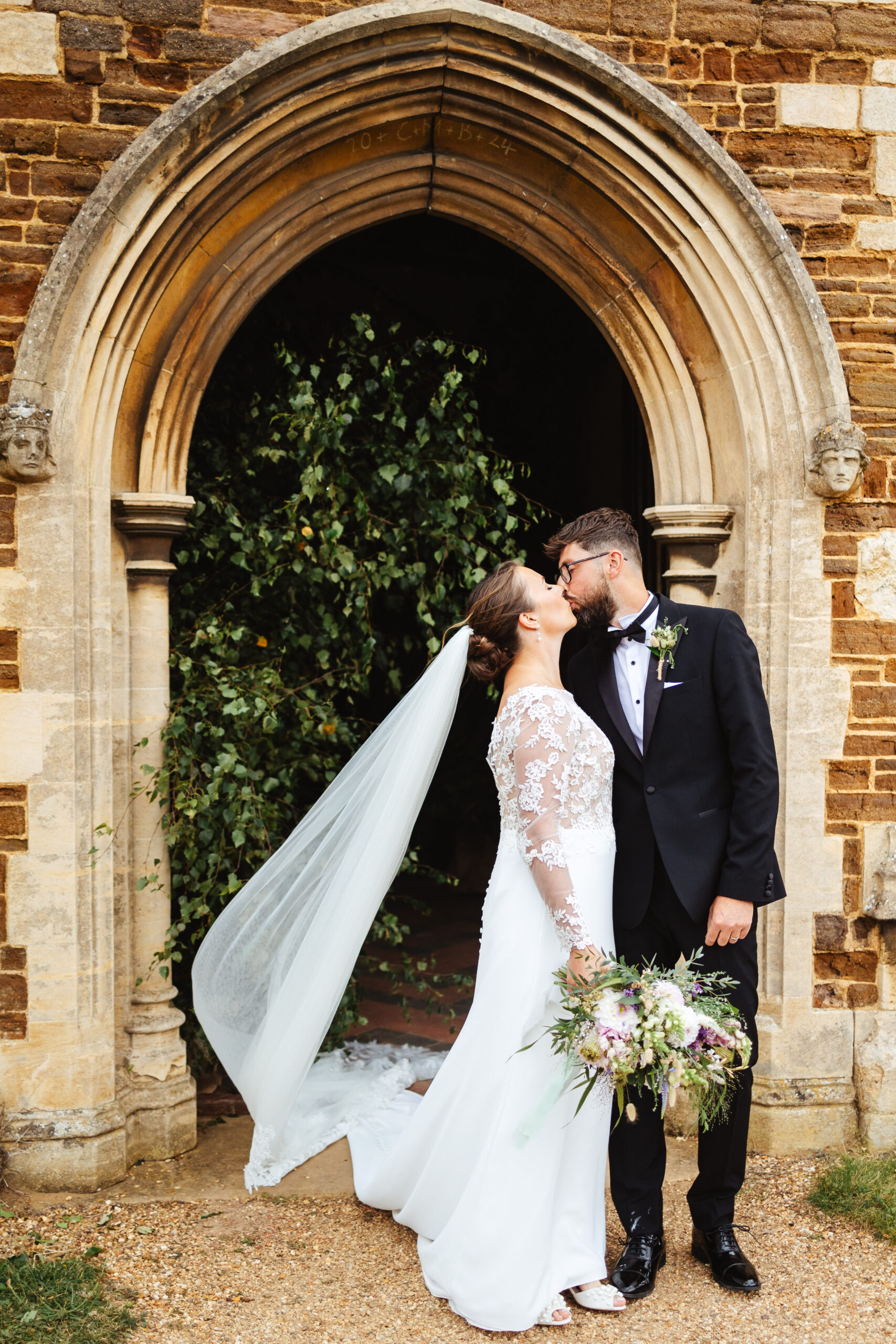 The bride and groom stood outside the church. They are kissing and the bride has her veil floating in the air. 