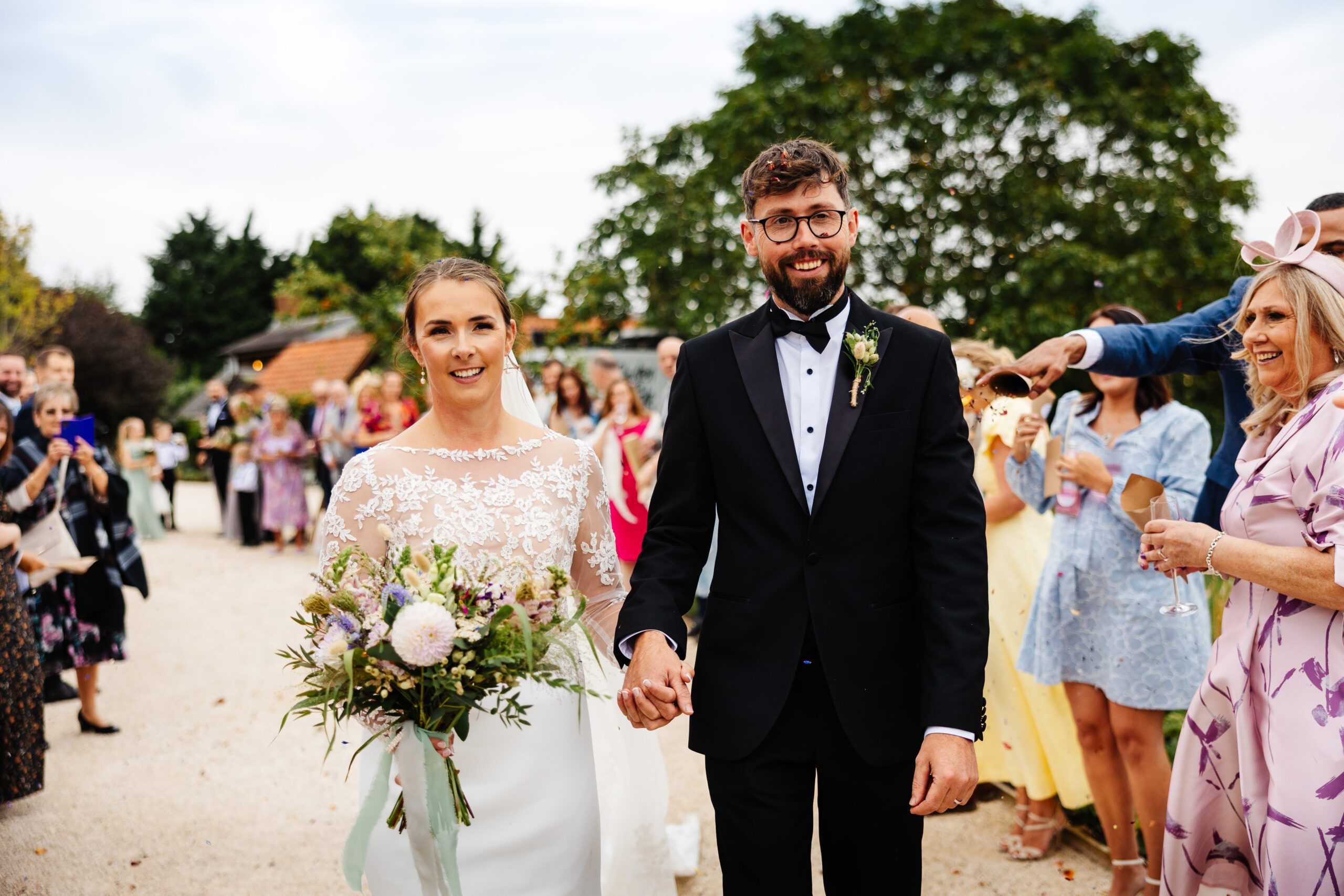 The bride and groom walking inbetween their guests. There is confetti floating in the air. The guests are smiling. 