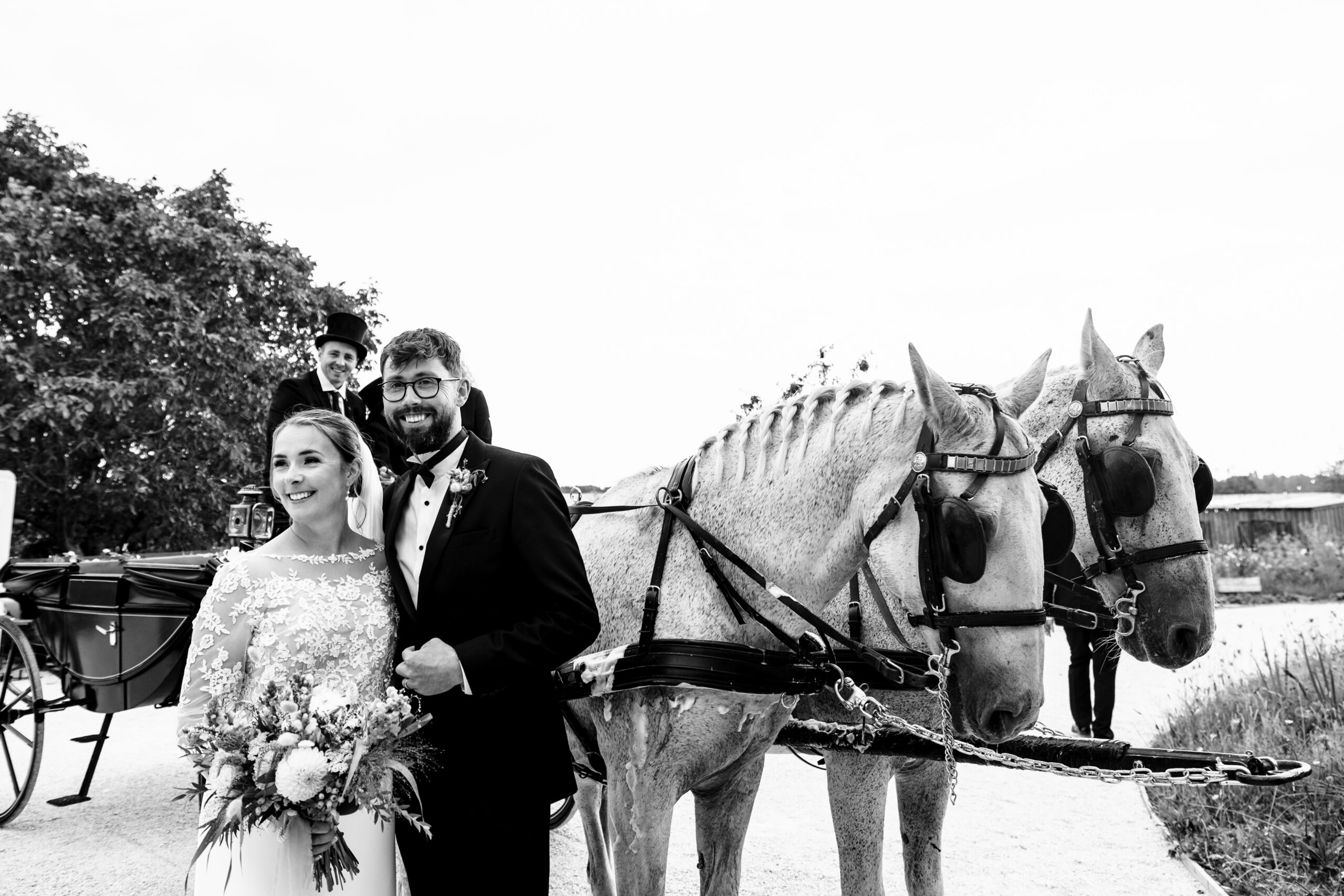 A black and white photo of the bride and groom in front of two horses. They are looking away from the camera and smiling.