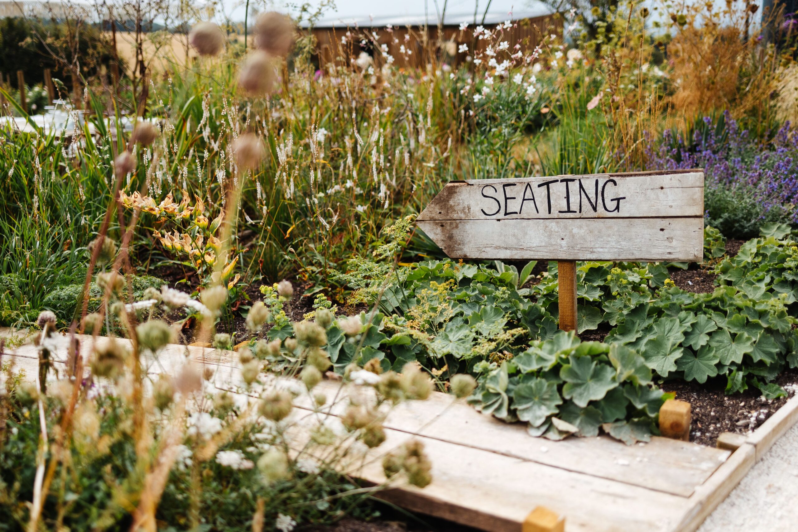 A handmade wooden sign that says Seating on it and an arrow pointing towards the gardens. The sign is dug in to lots of greenery and garden.