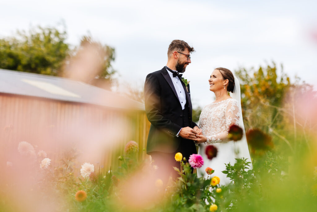 bride and groom stand looking at one another in the background. The foreground is full of colourful dahlias.