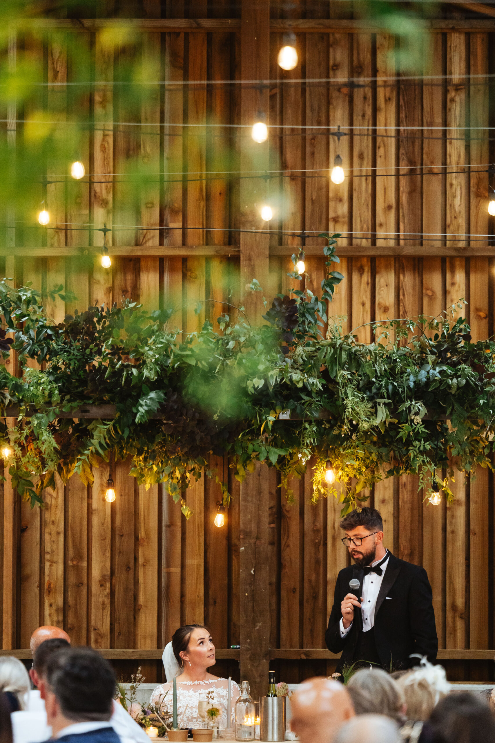 A portrait shot of the bride and groom. The groom has a microphone and is giving a speech. The bride is looking at the groom and looks emotionally happy. 