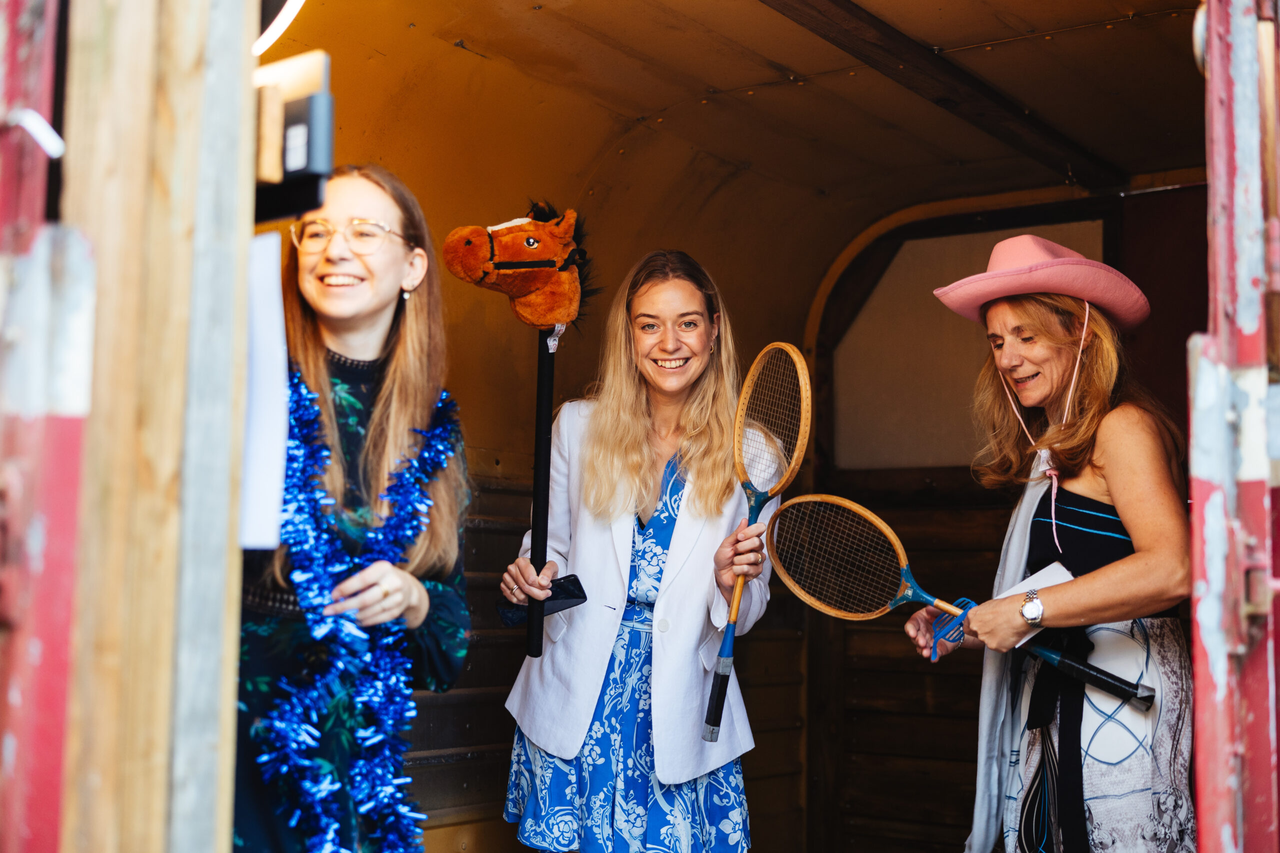 Three guests in the horsebox photobooth. They are smiling and laughing and holding props such as cowboy hats, tennis rackets and a horse on a stick. They are having a good time.