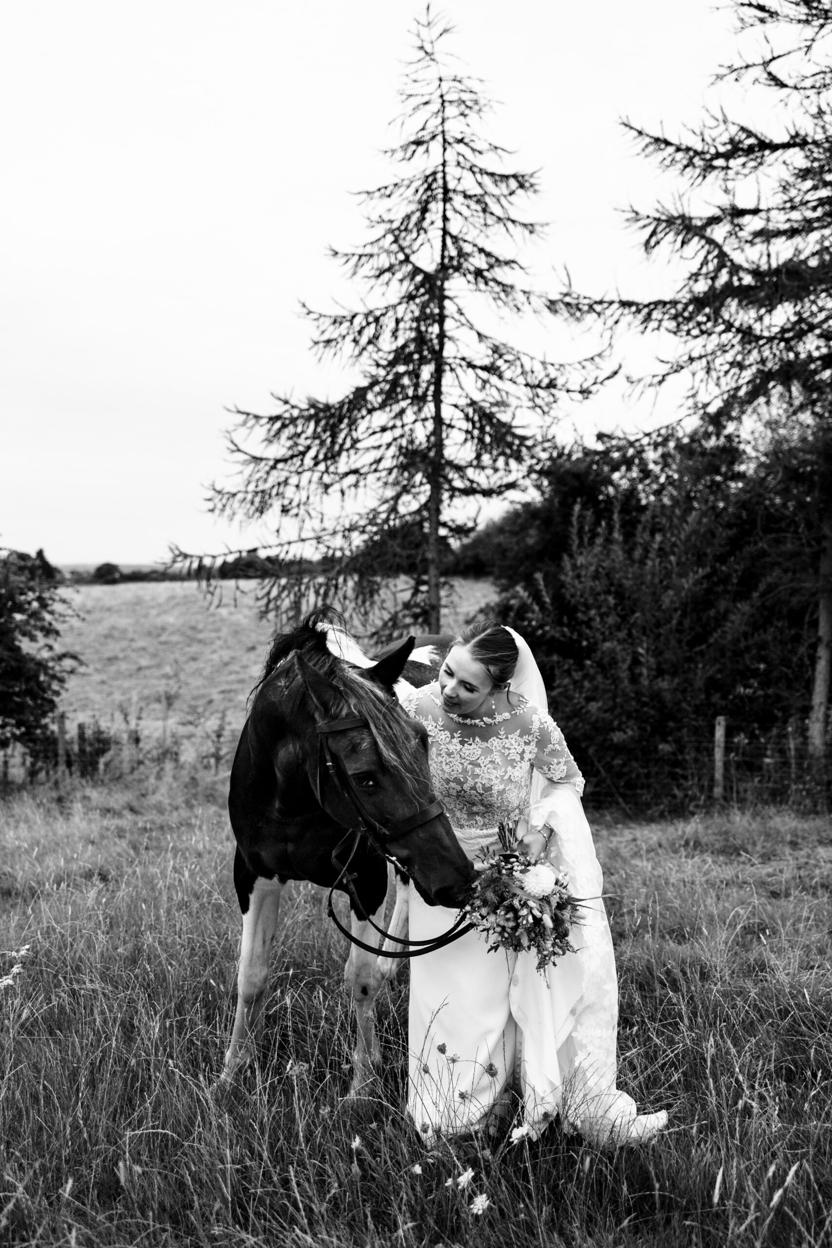 A black and white image of the bride. She is standing next to her horse and is looking at it and stroking it.