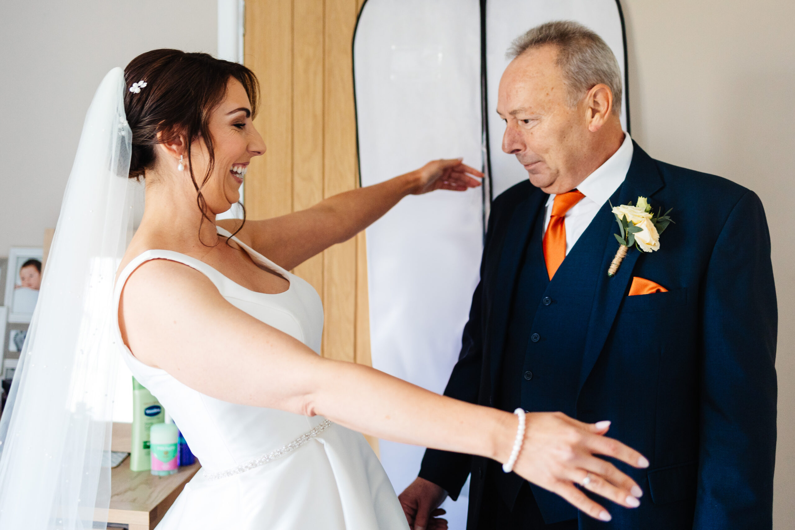 The father of the bride seeing the bride for the first time. He is looking at her and he is emotional. The bride is holding her arms out for a hug and is smiling.