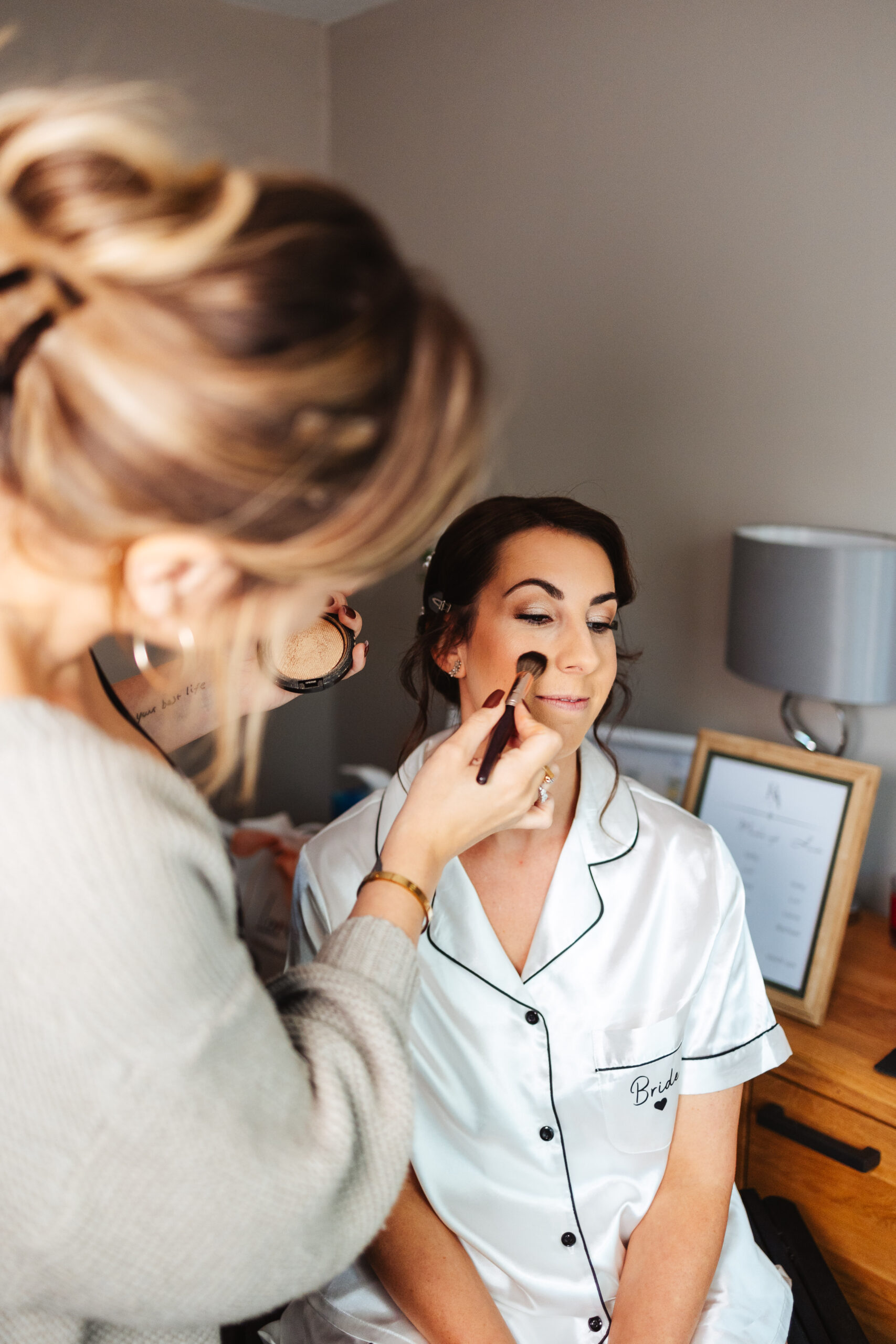 The bride having her makeup done. She is wearing silk white pyjamas with a black edging. You can see the back of the make up artist and she is putting a blusher on the bride. The bride is smiling.