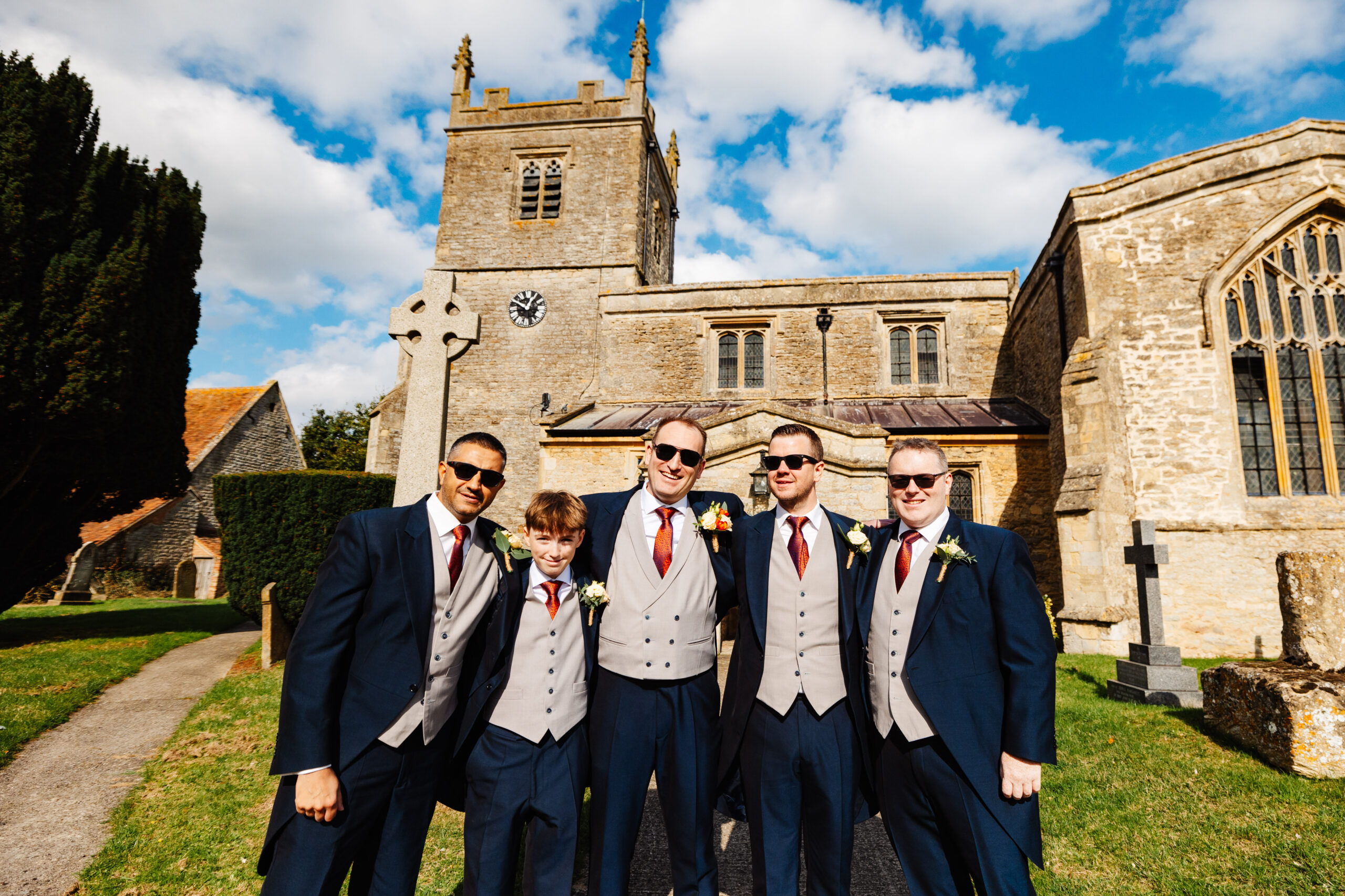 The groom and his groomsman stood outside the church. They are wearing sunglasses and smiling.