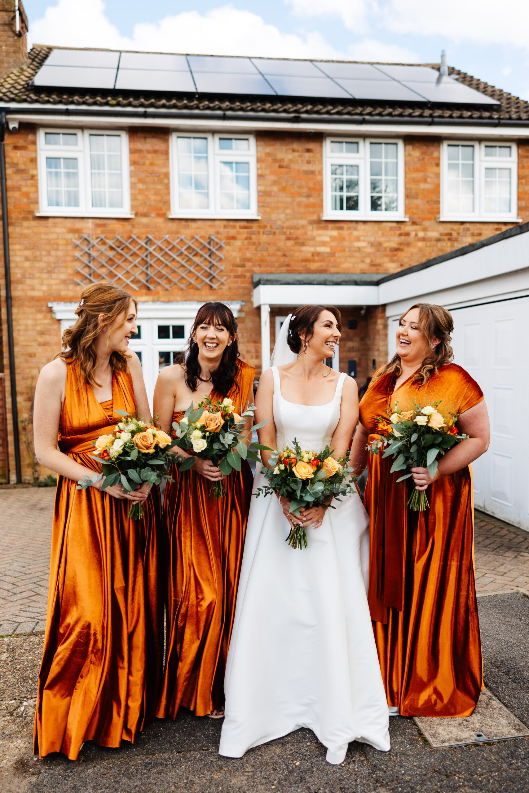 The bride and her bridesmaids ready for the wedding. They are stood outside a house. The bridesmaids are wearing rustic orange dresses. They are all looking at each other and smiling. They are holding bouquets of flowers and the flowers are a mixture of orange and yellow roses and Eucalyptus.