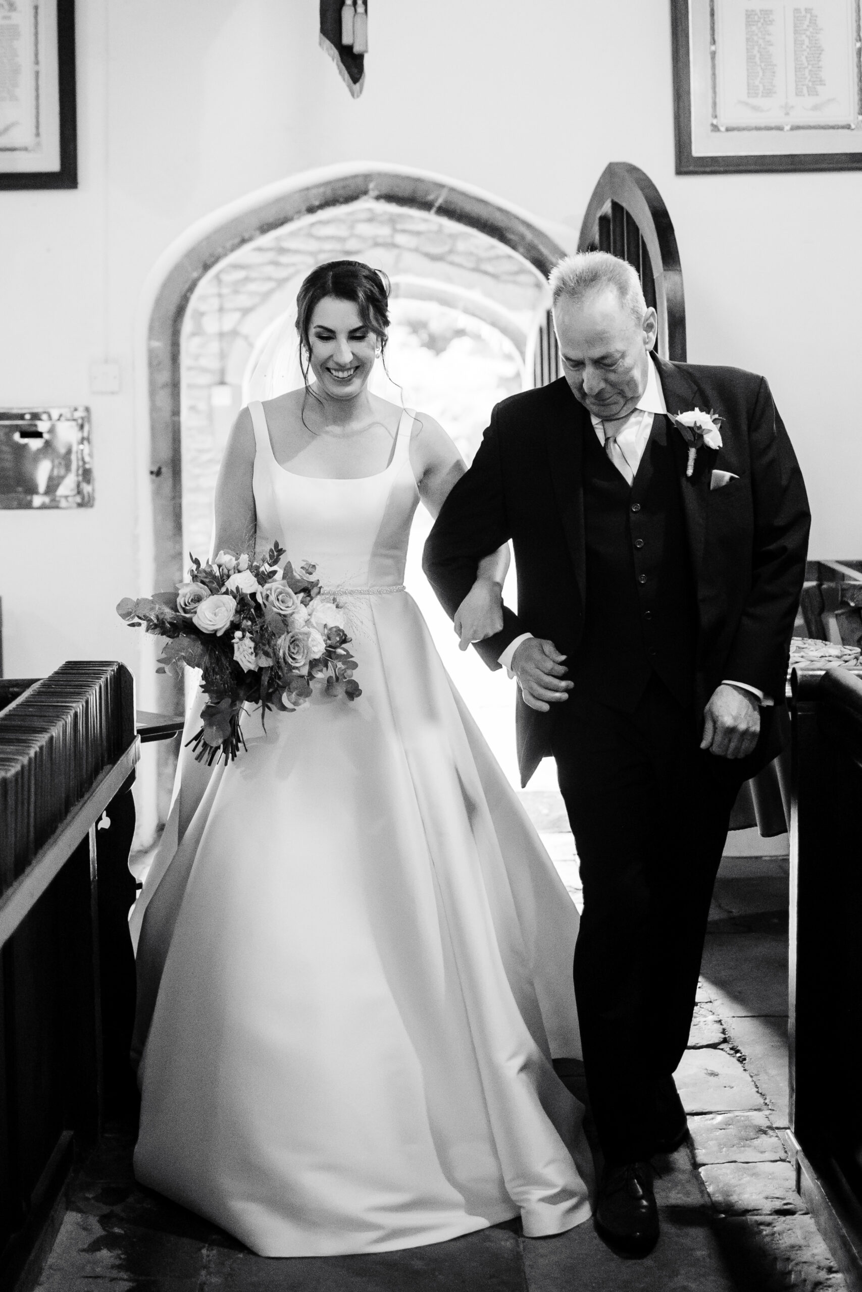 The father of the bride walking the bride down the aisle. The father is crying and the bride is smiling. They are arm in arm walking down the aisle in the church.