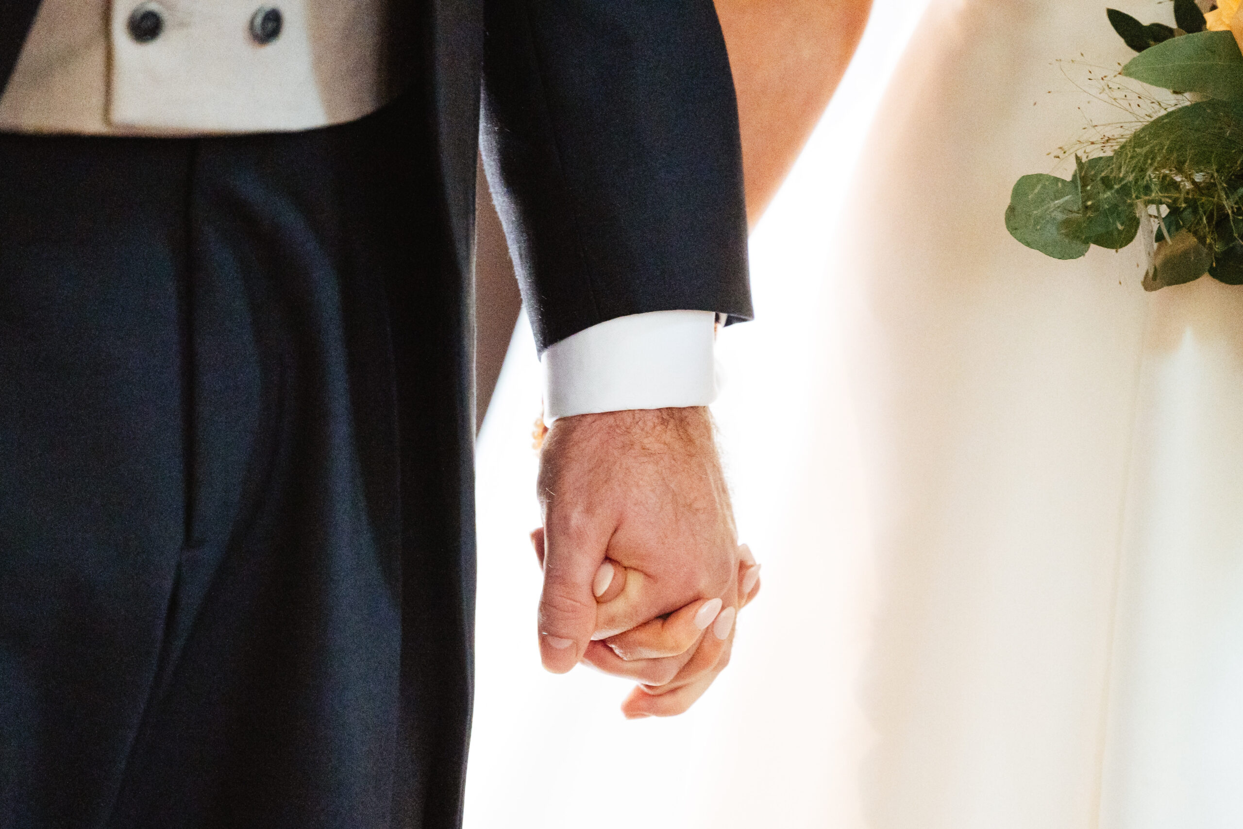 A close up shot of the bride and groom's hands. They are holding hands. The bride's dress and the groom's bottom of the waistcoat are in shot.