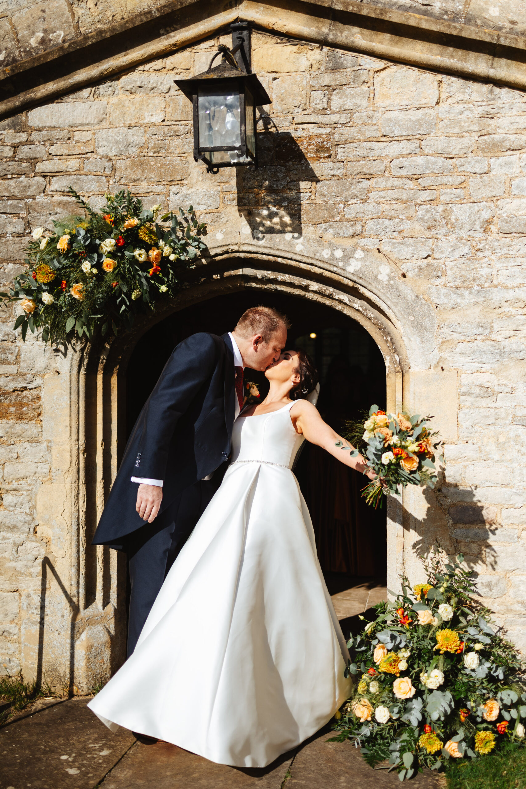 The bride and groom outside of the church sharing a kiss. The bride is leaning back and is holding her flowers out to the side. You can see the beautiful stone church in the background.