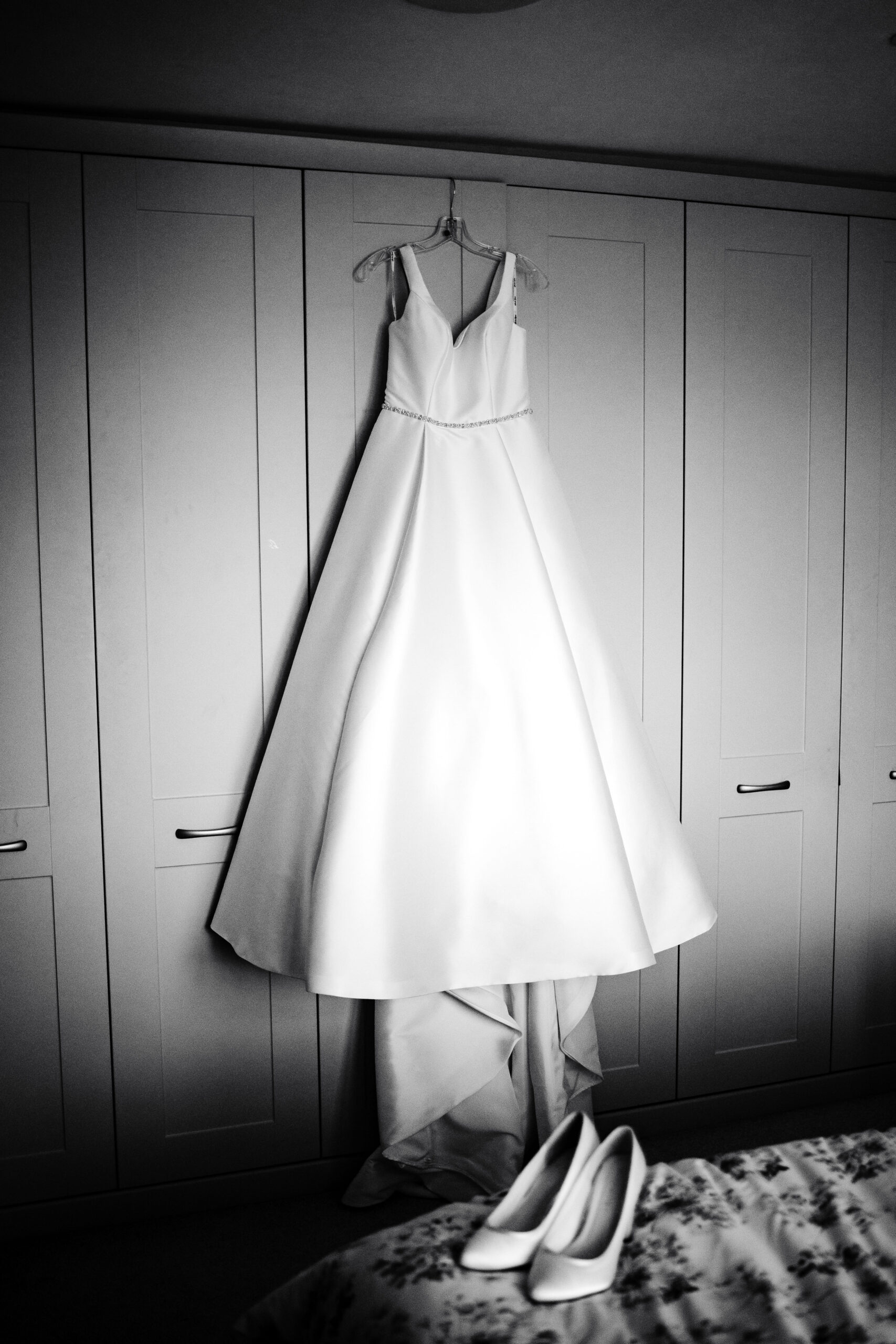 A black and white image of a wedding dress. The dress is hung up on a wardobe. It is white with a little diamante beaded waist belt.