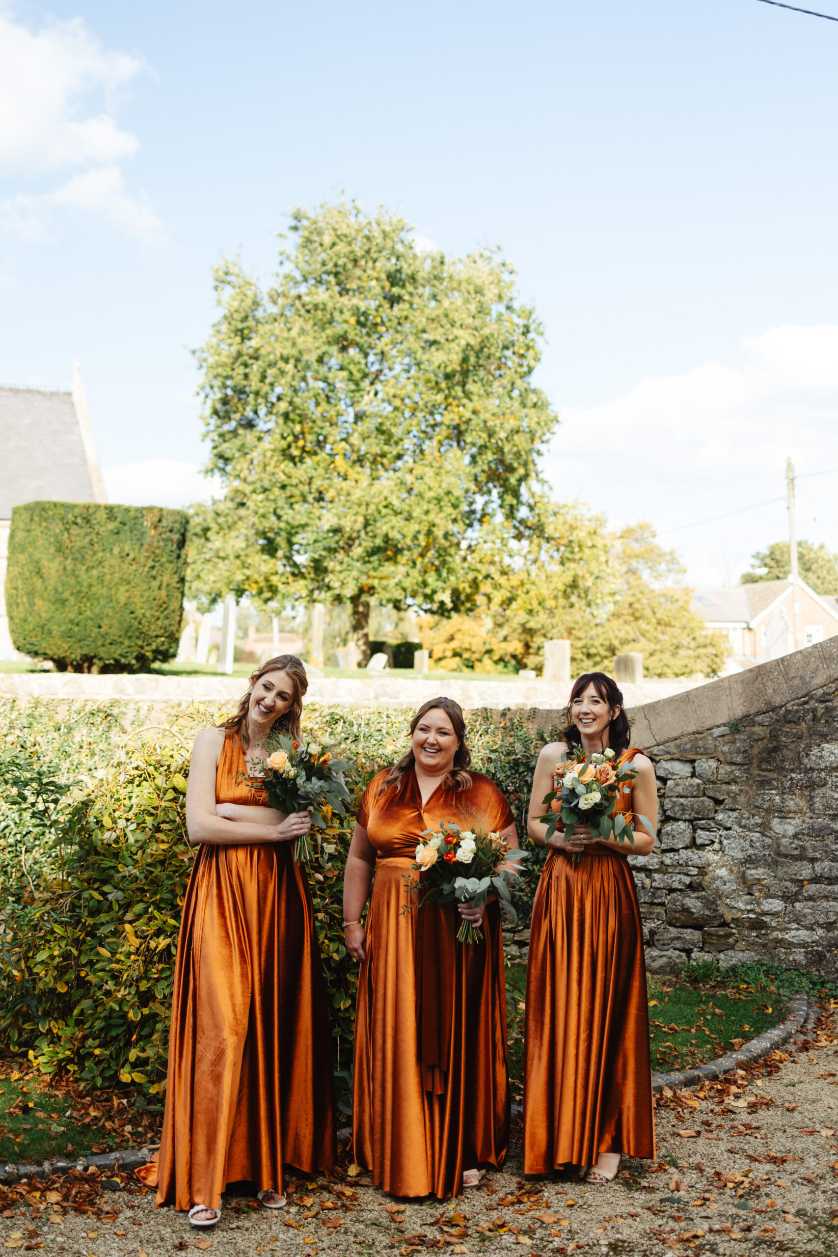 The bridesmaids standing looking at the bride and groom. They are in rustic orange, long silky dresses.