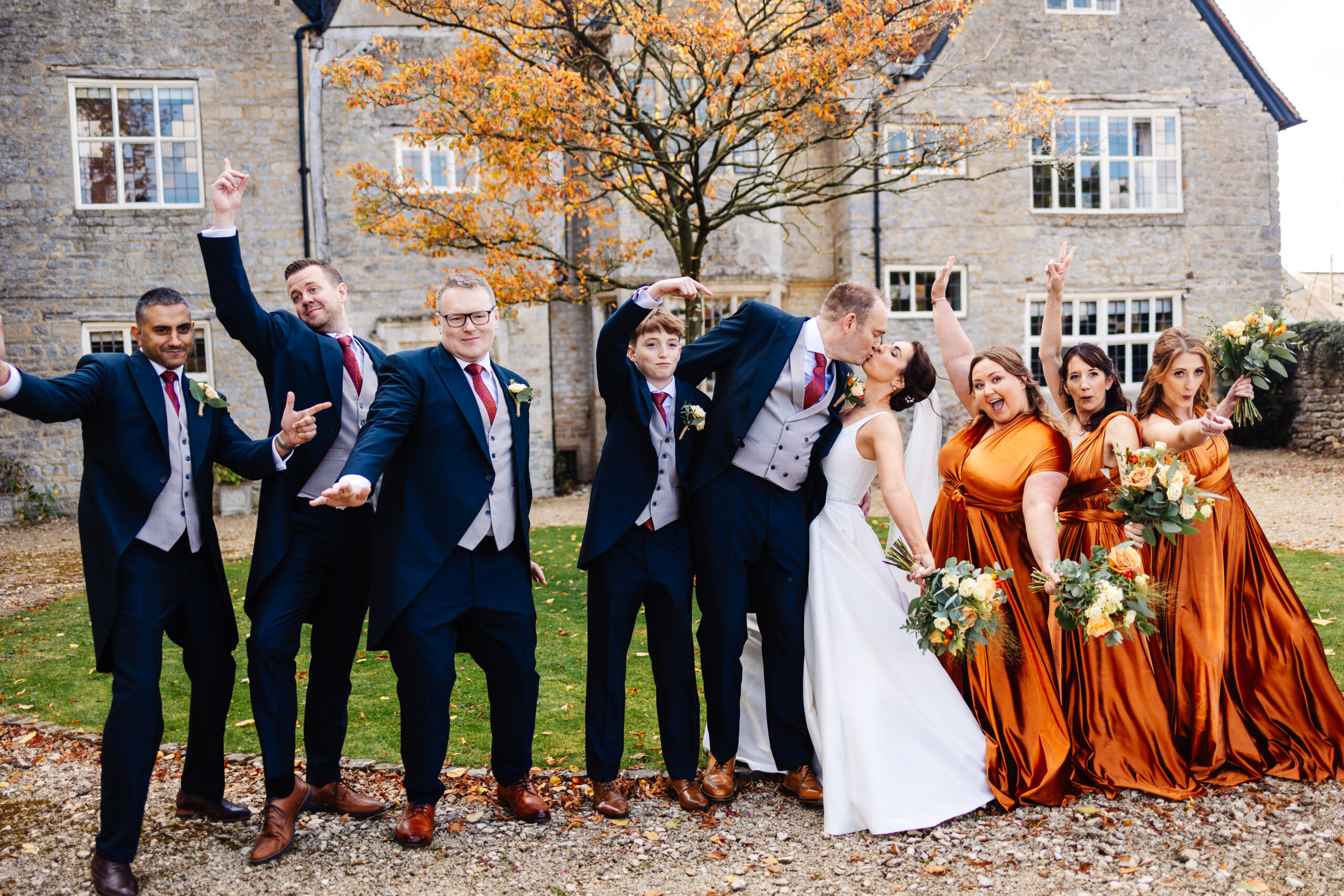 A group shot of the bridal squad. They are all having fun and throwing their hands in the air whilst the bride and groom kiss.