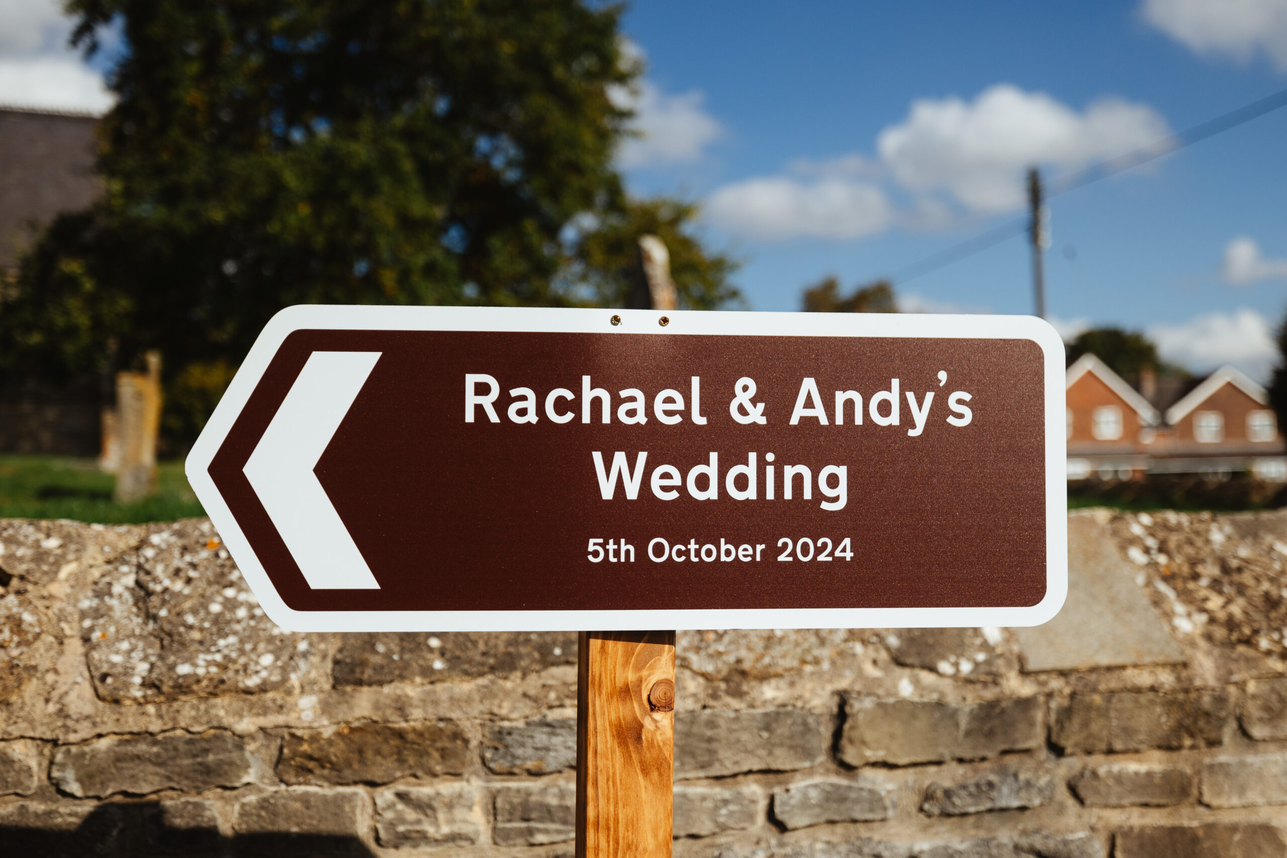 A brown sign that says Rachael & Andy's Wedding. 5th October 2024. This is in front of the church where they will get married, signposting guests to the right location.