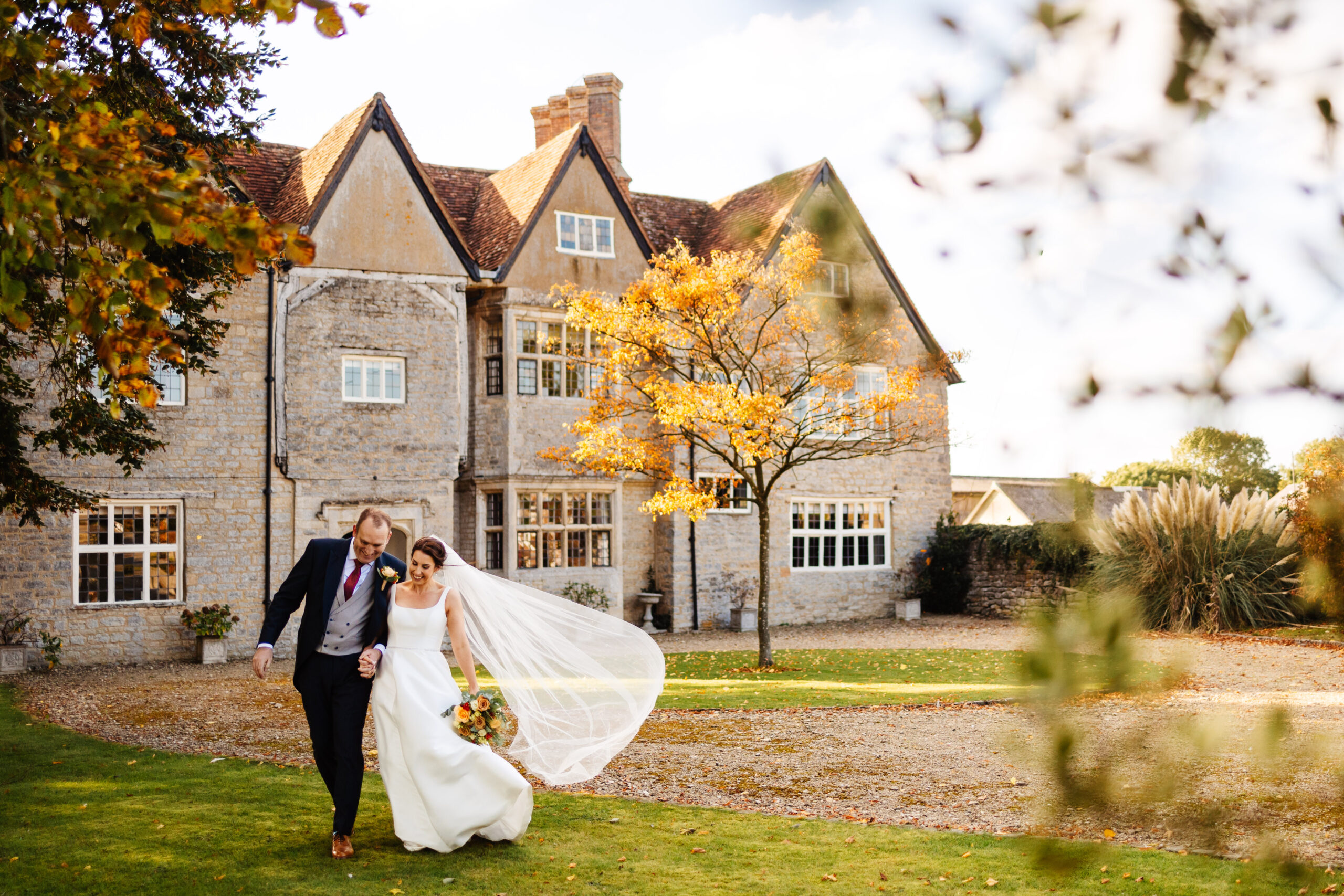 bride and groom laughing together in front of manor farm barn manor house. The leaves on the trees are yellow and the sun is low during autumn