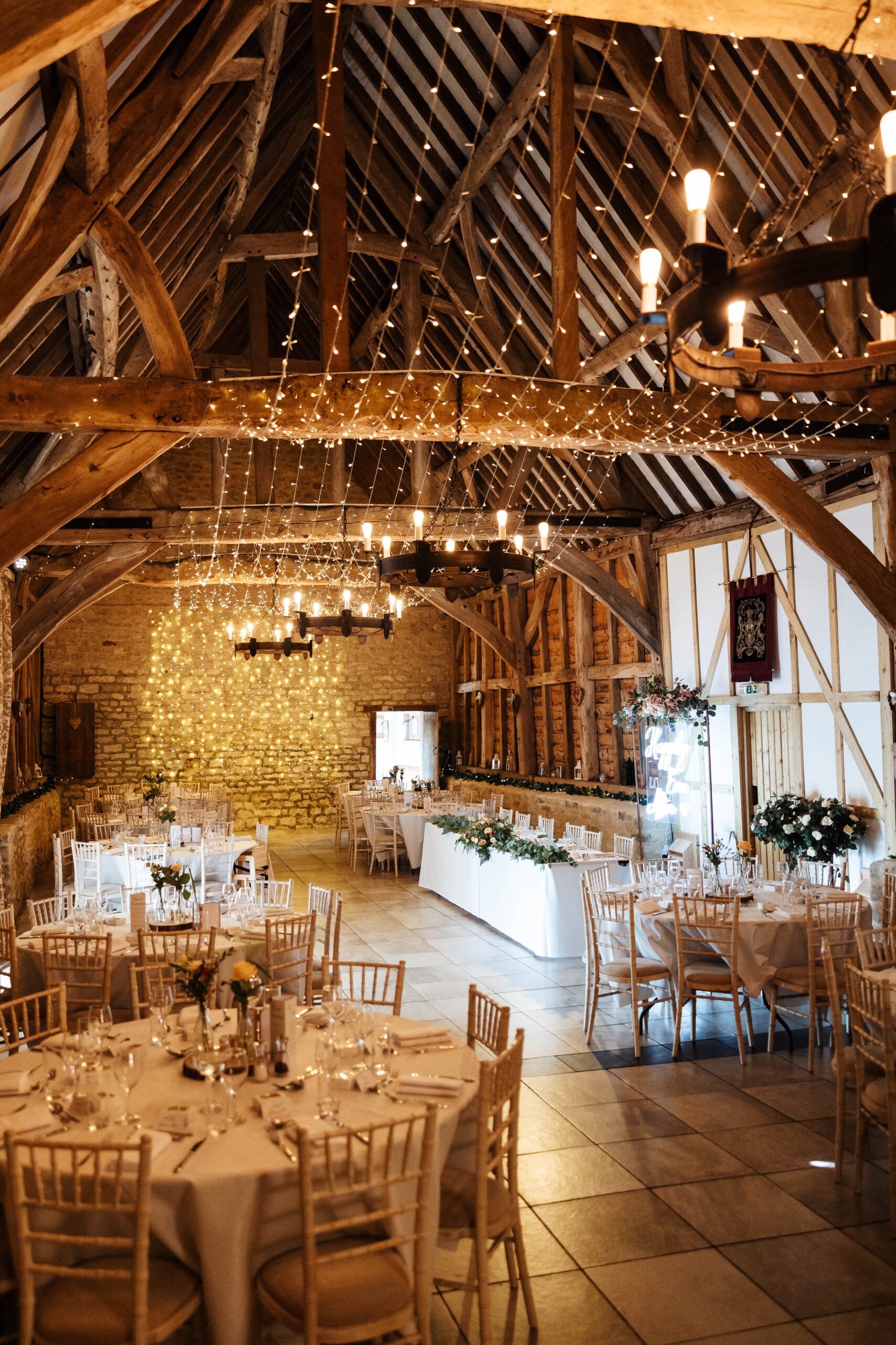 The venue. It is an old barn with old beams and you can see round tables as well as a rectangular top table. There are flowers on the top table and the chairs are wooden. There are fairy lights in the beams and on the back wall.