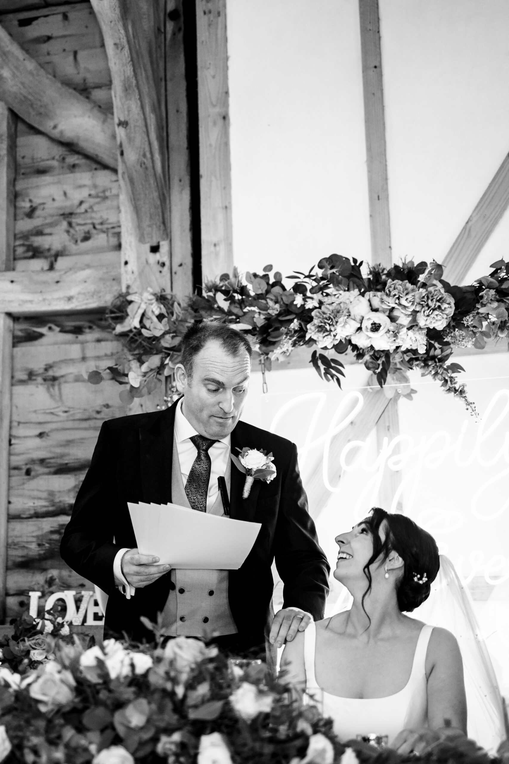 The groom giving a speech to his bride. They are looking at each other and smiling. The groom has his speech in one hand and has his other hand on her shoulder.