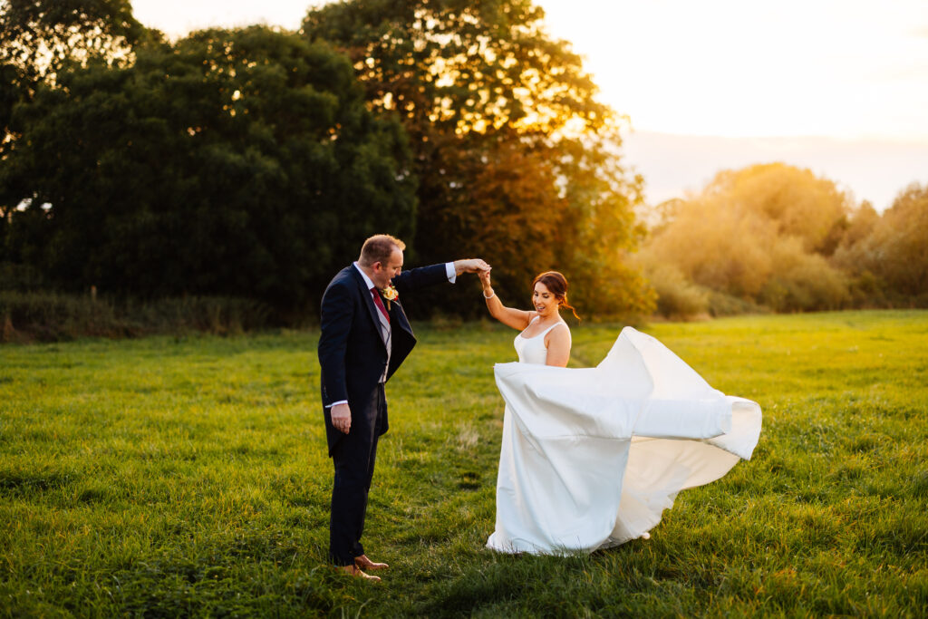 bride and groom during golden hour, low sun shining across green fields at manor farm barn. The groom is twirling the bride around and her dress trails behind her. 