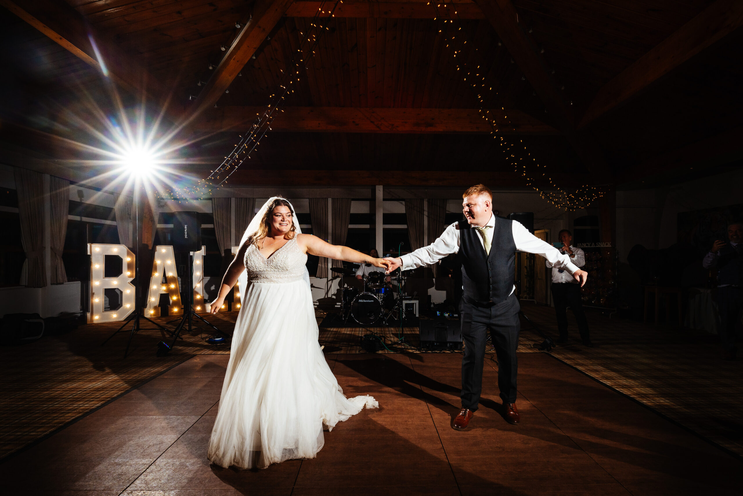 The bride and groom alone on the dance floor. You can see light up letters in the background that say BALL. The couple are holding hands and are about to dance. They are smiling.