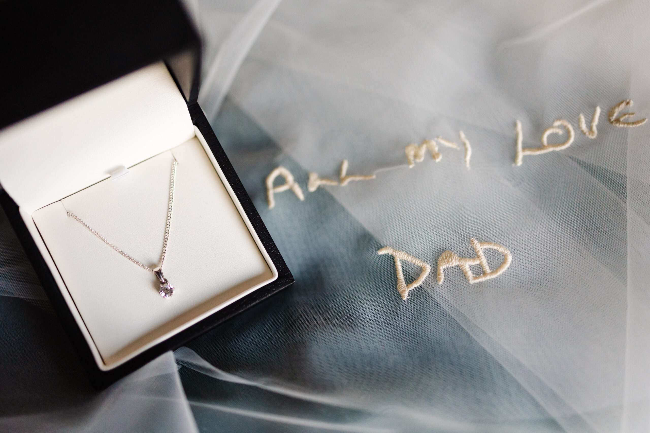 A silver necklace with a small diamond pendant which is in a box and the box is on top of a white veil which has the words 'All My Love, Dad' embroidered on it.