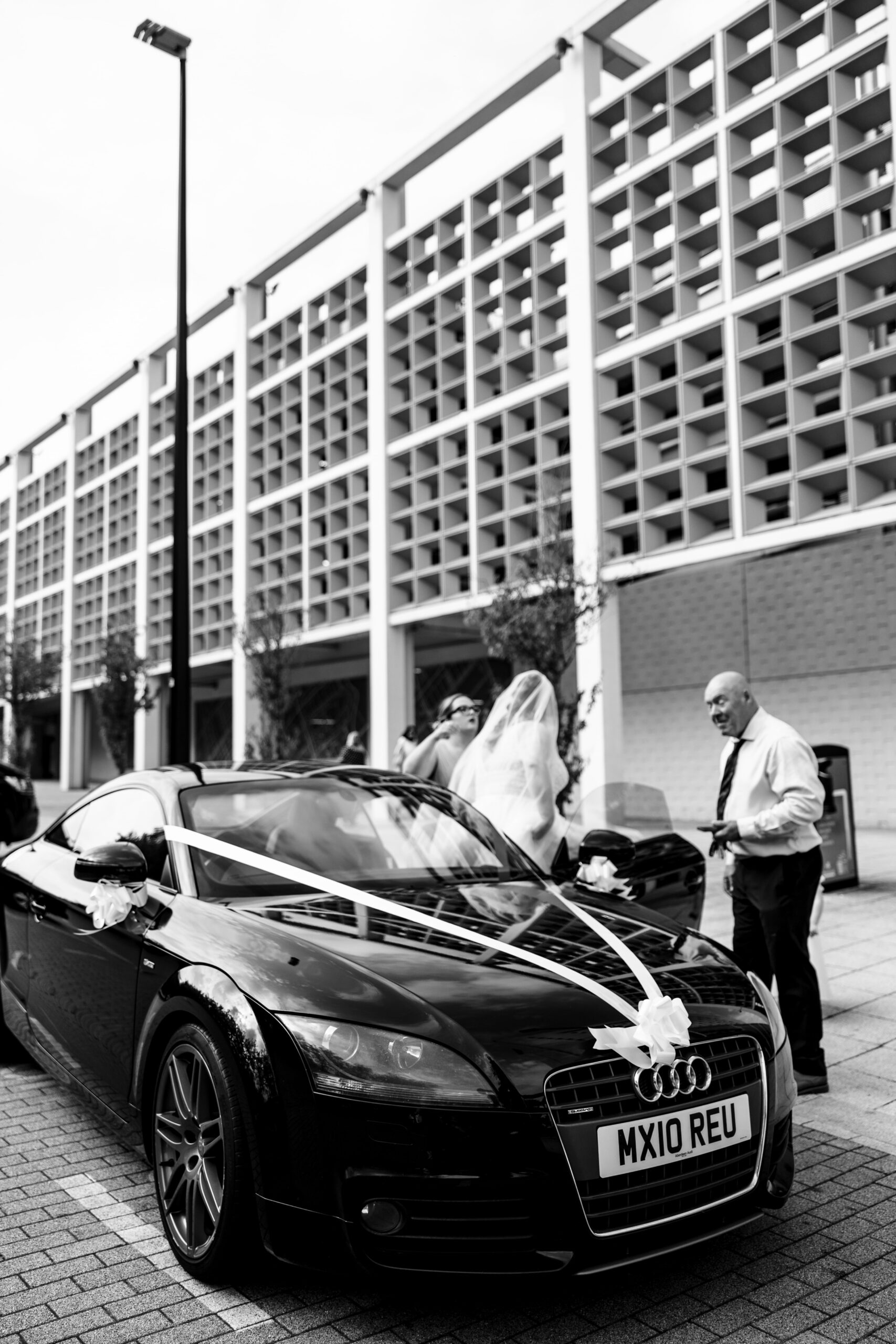 A sleek, black convertable audi with a white ribbon on the front. The bride's father is helping the bride get in the car.