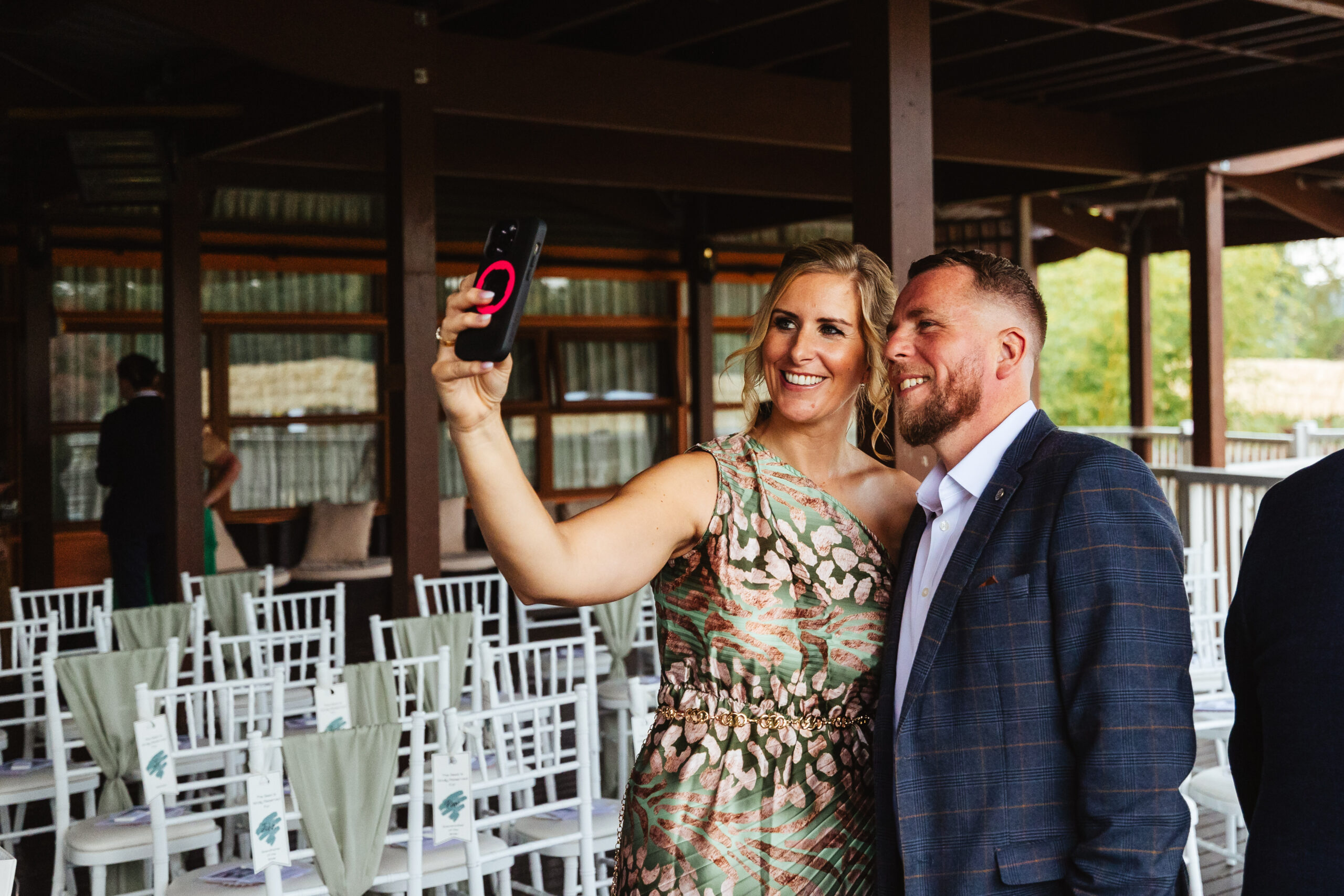 Two wedding guests smiling for a selfie at the wedding venue. You can see chairs in the background that have a light green ribbon on; matching the colour of the groom's ties.