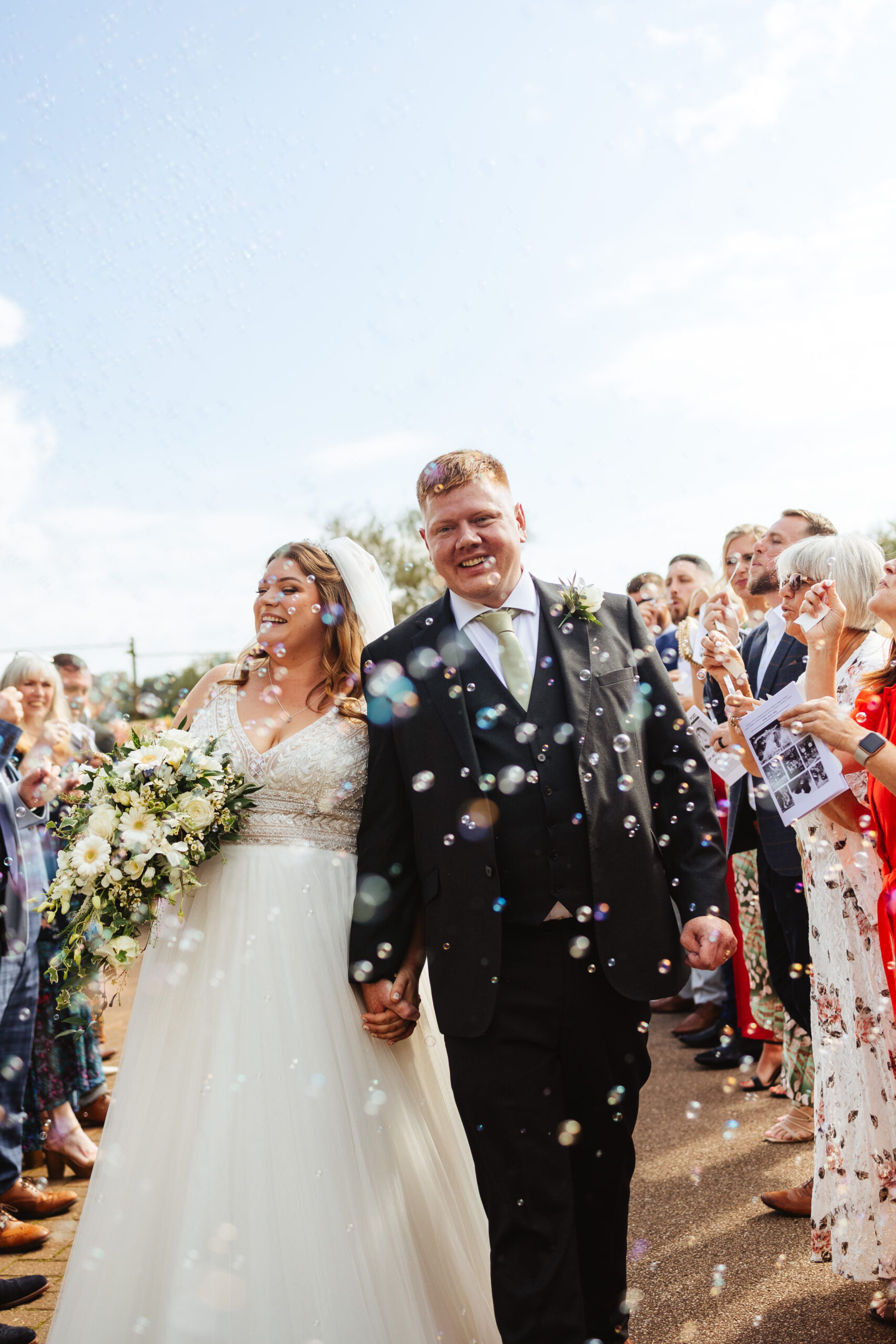 The bride and groom walking through a line of their guests. The guests are blowing bubbles as the couple walk. You can see lots of bubbles surrounding the couple and the guests in the background blowing them. The bride and groom are smiling.