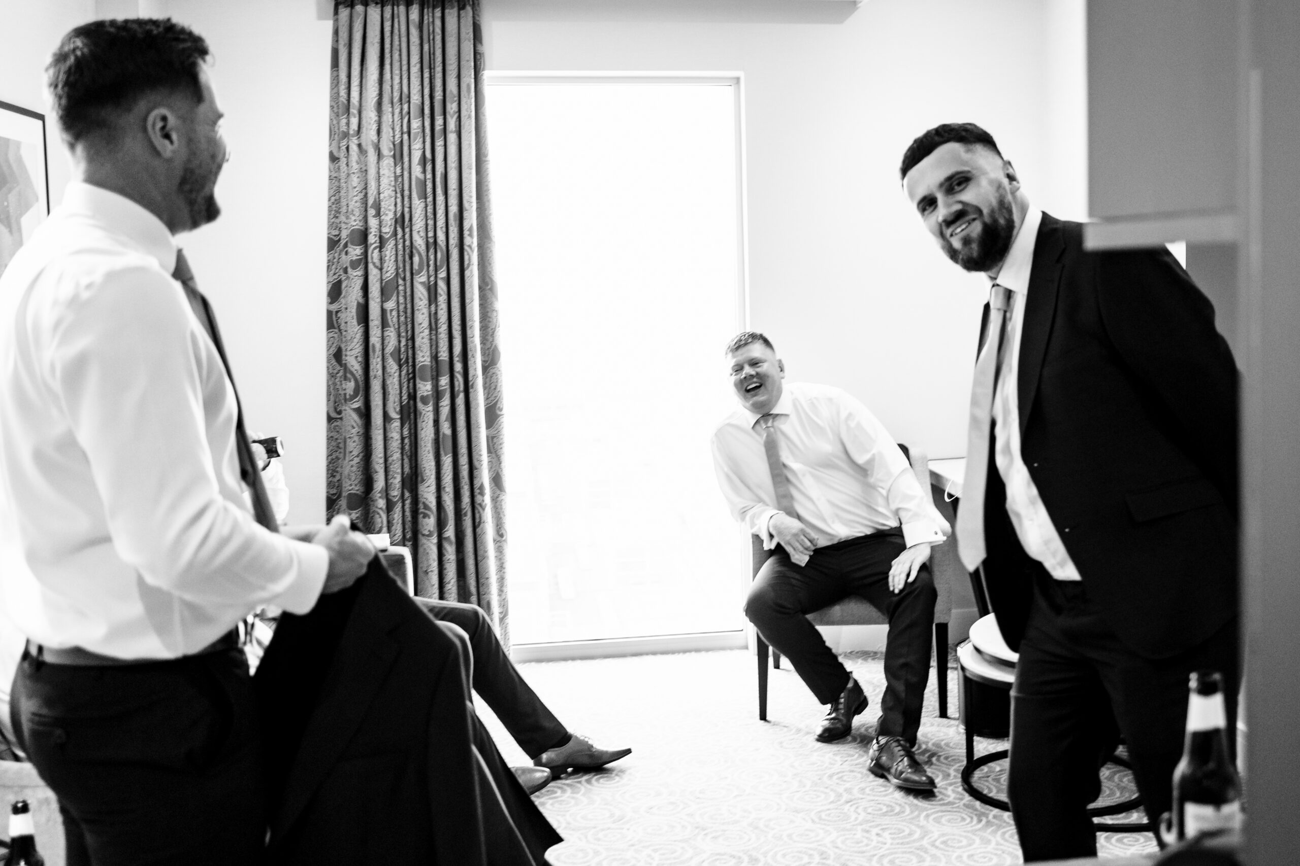 A black and white image of the grooms team getting ready. Two men are standing up, wearing white shirts and black blazers and the groom is sat down on a chair looking relaxed.