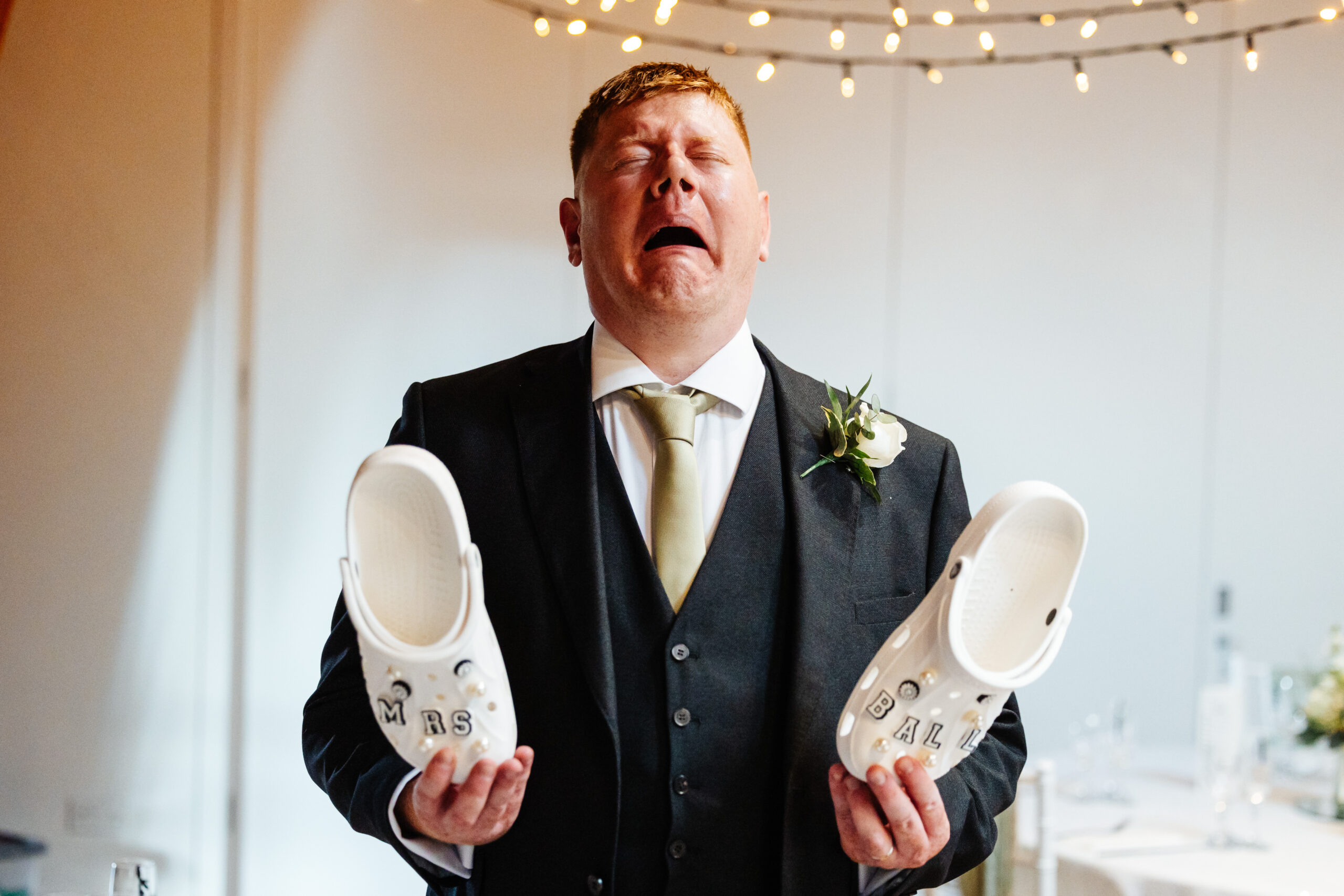 The groom pulling a disgusted face. He is holding a pair of white crocs that say Mrs Ball on them.