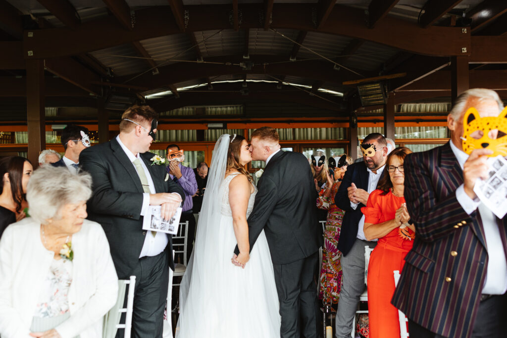 bride and groom walking back down the aisle hand in hand, having a kiss. Their wedding guests look directly at them and clap or smile. 