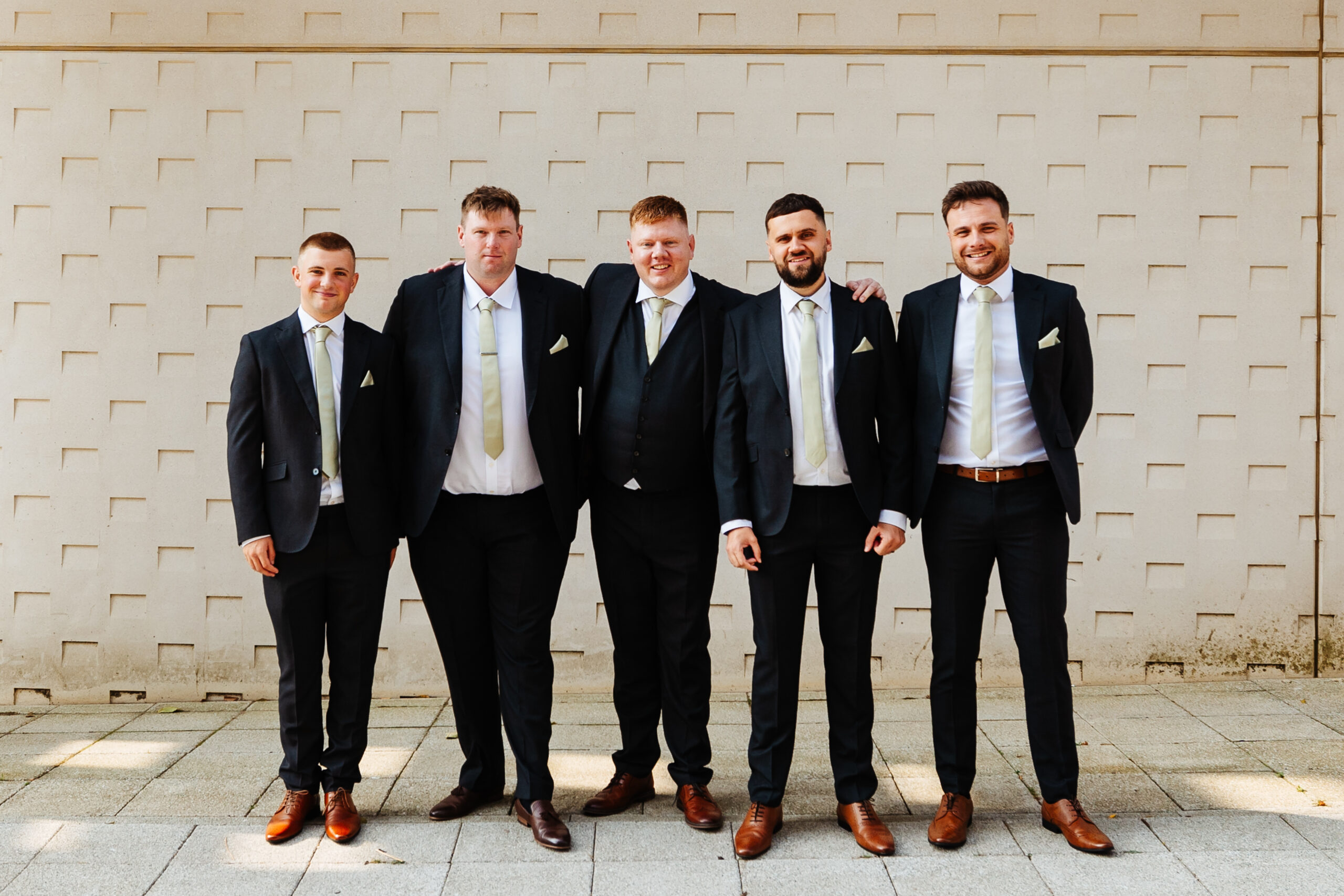 The groomsmen stood outside a white, textured wall. They are wearing black blazers, white ties and pale green ties. They are wearing brown shoes and all looking at the camera smiling.