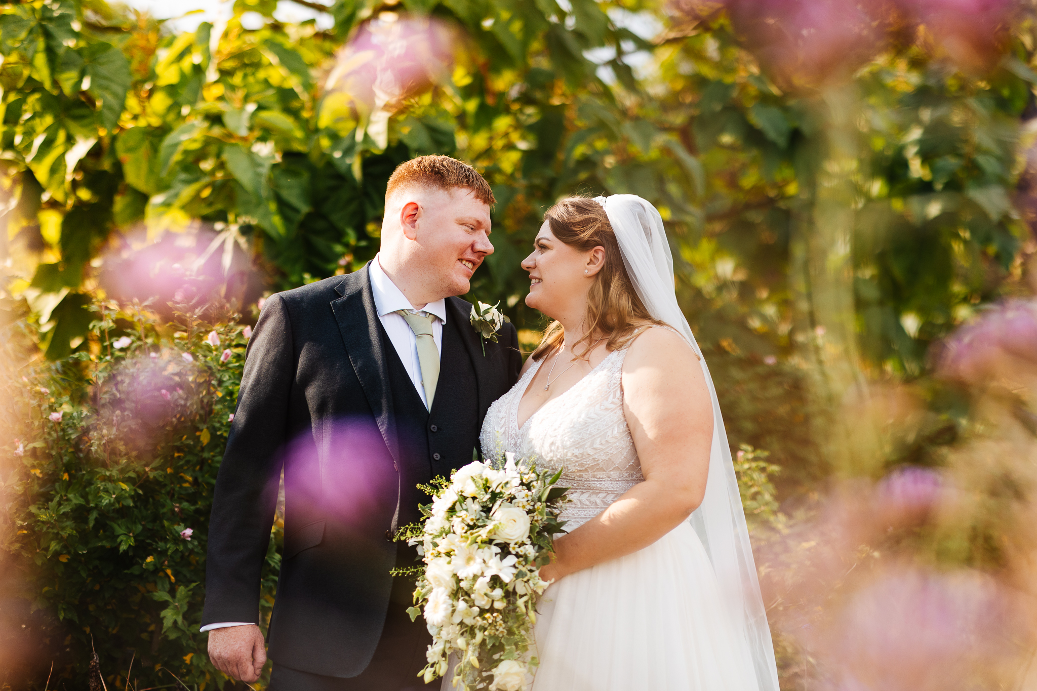 bride and groom standing close together looking at one another. They're surrounded by green foliage dusted with pink flowers, at woburn safari park