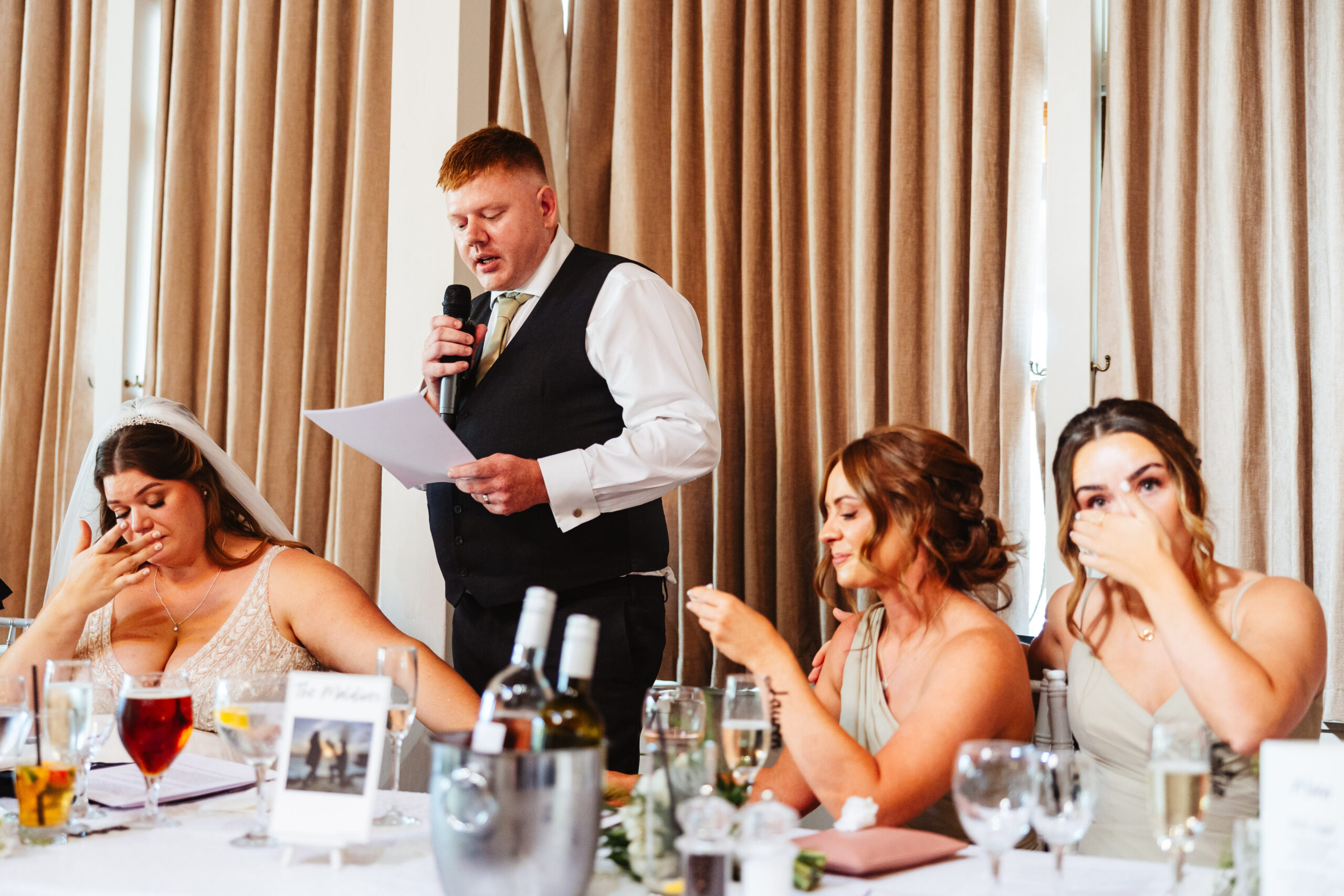 The groom giving a speech. He has taken his jacket off and is wearing a waistcoat. The two bridesmaids are wiping their eyes from tears and the bride is also very emotional.

The groom is holding a piece of paper in one hand (his speech) and a microphone in the other.